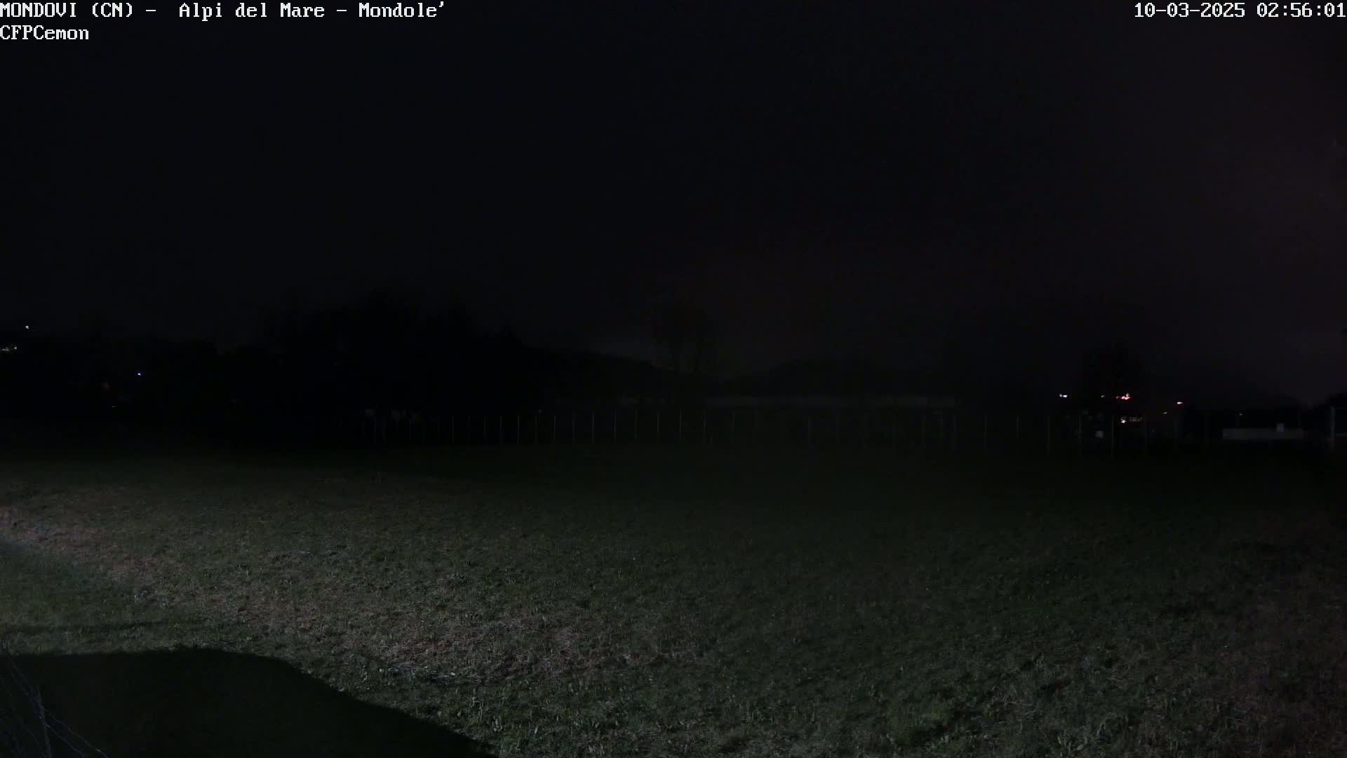 A dark, nighttime view of a grassy field with a fence and distant, faintly lit structures under a mostly clear sky.