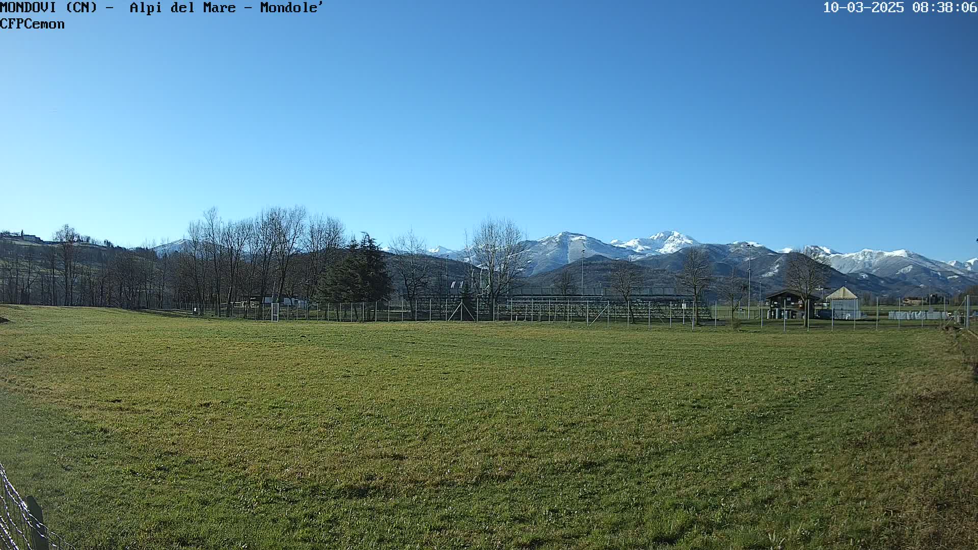 A grassy field with a fenced-in area and bleachers sits in the foreground, under a clear blue sky, with snow-capped mountains in the background.