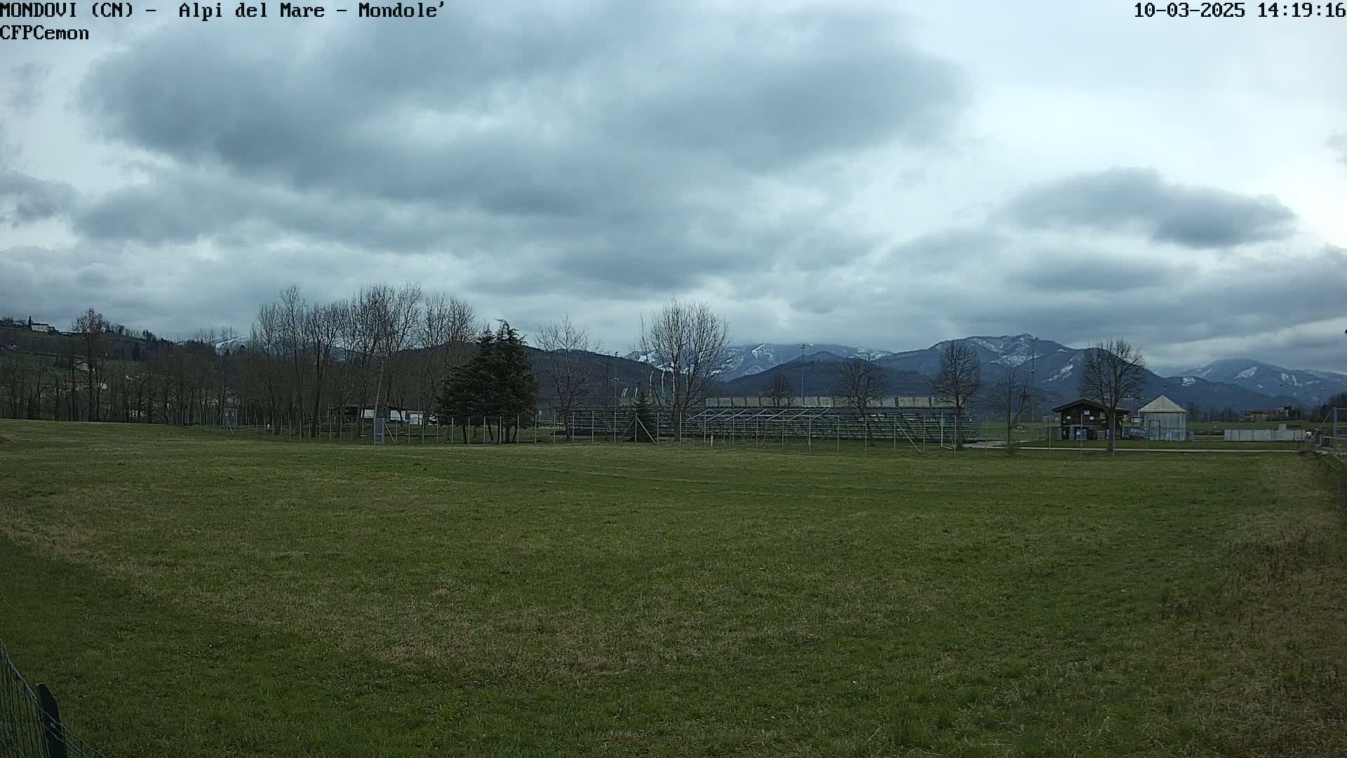A grassy field stretches out before empty bleachers, beyond which are bare winter trees and snow-capped mountains under a cloudy sky.