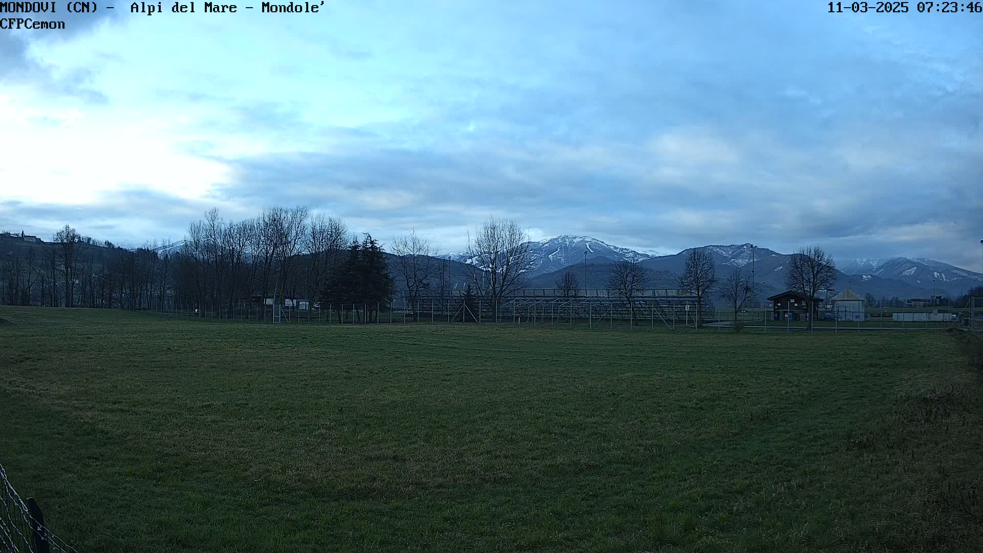 A grassy field with a fence and bleachers in the foreground stretches towards snow-capped mountains under a mostly cloudy sky.