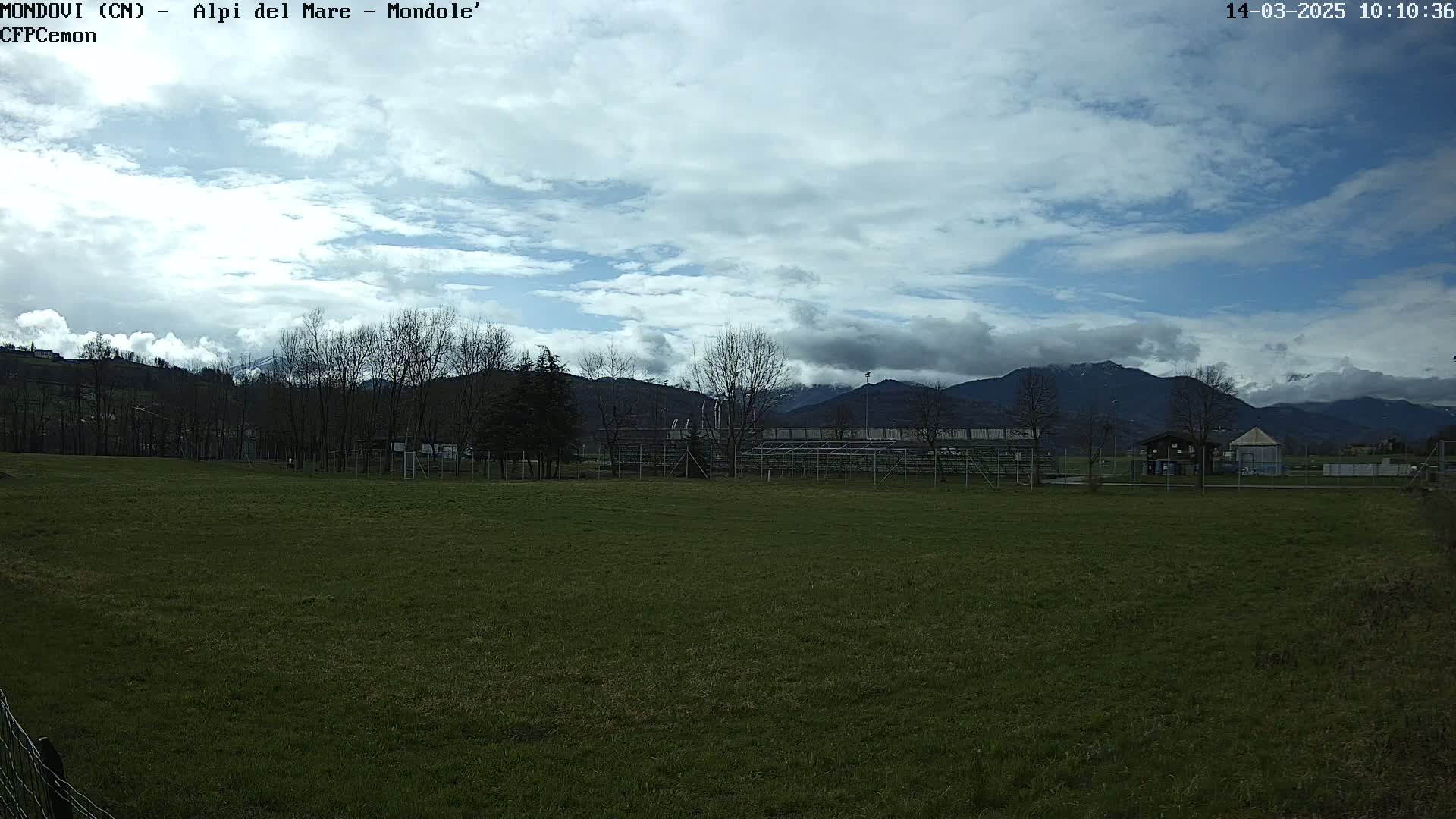A partly cloudy day reveals a grassy field with a small structure and bleachers in the middle ground, beyond which are bare trees and snow-capped mountains in the background.