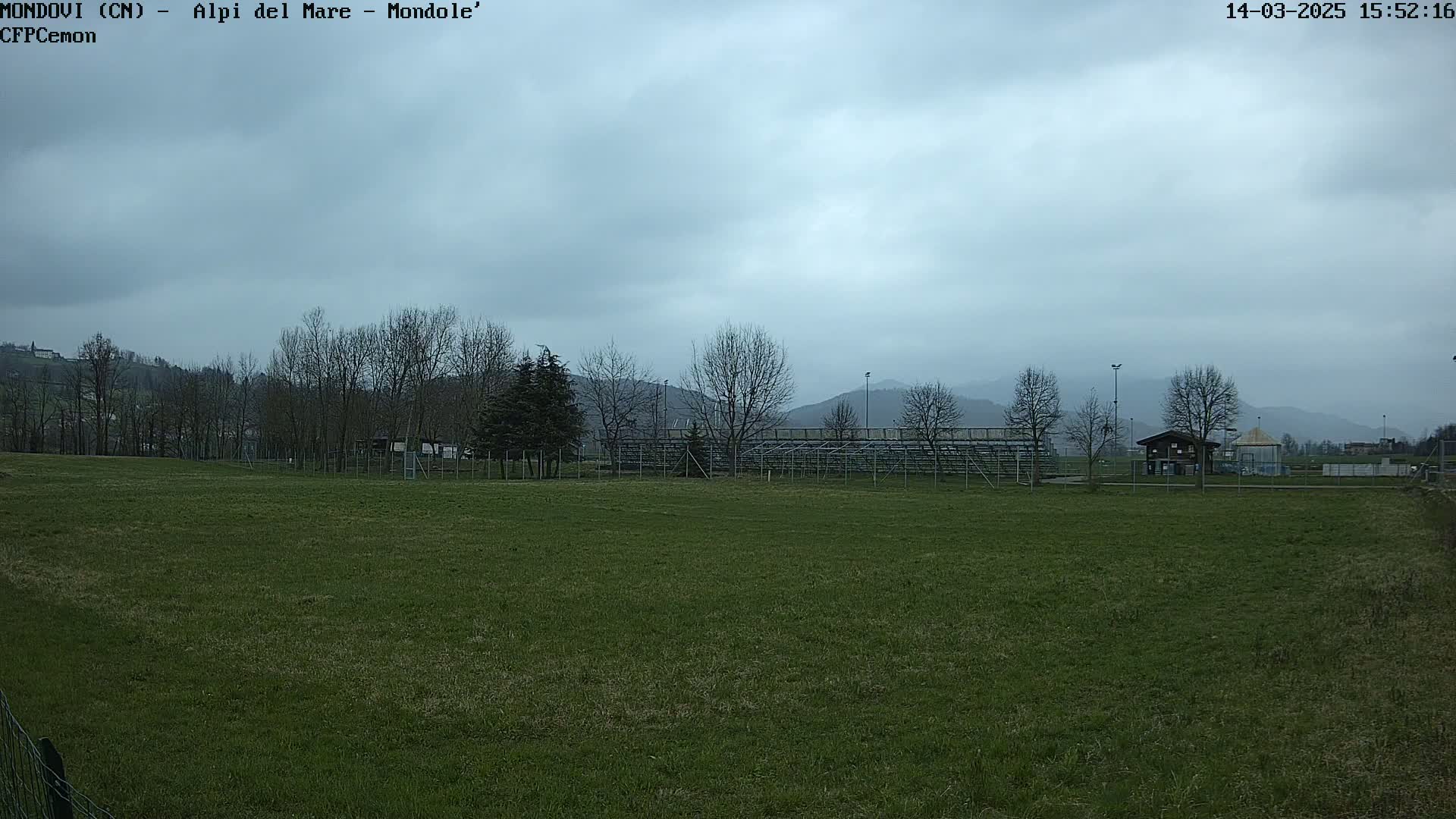 A grassy field stretches out under an overcast sky, with bare trees, bleachers, and a small building visible in the mid-ground, and distant hills in the background.