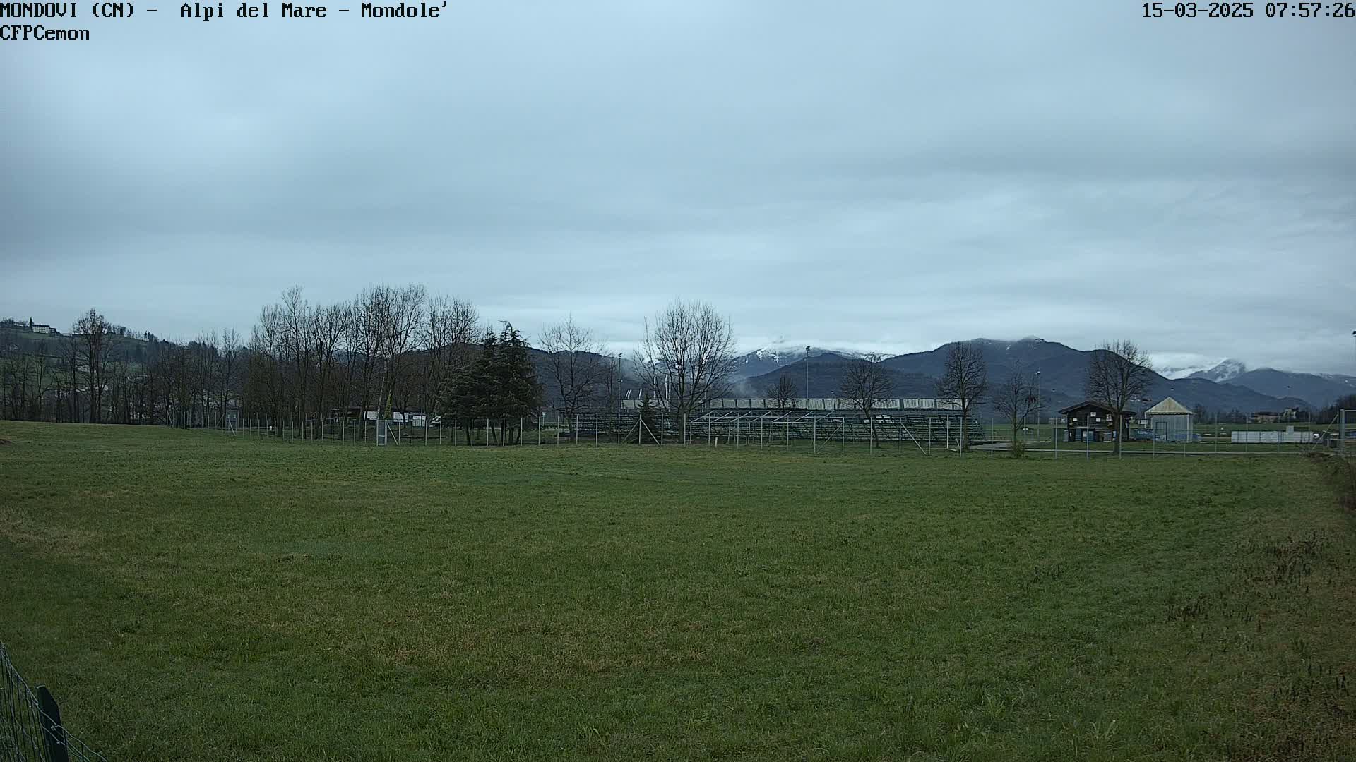 A grassy field with a fence and bleachers is in the foreground, behind which are bare trees and snow-capped mountains under a cloudy sky.