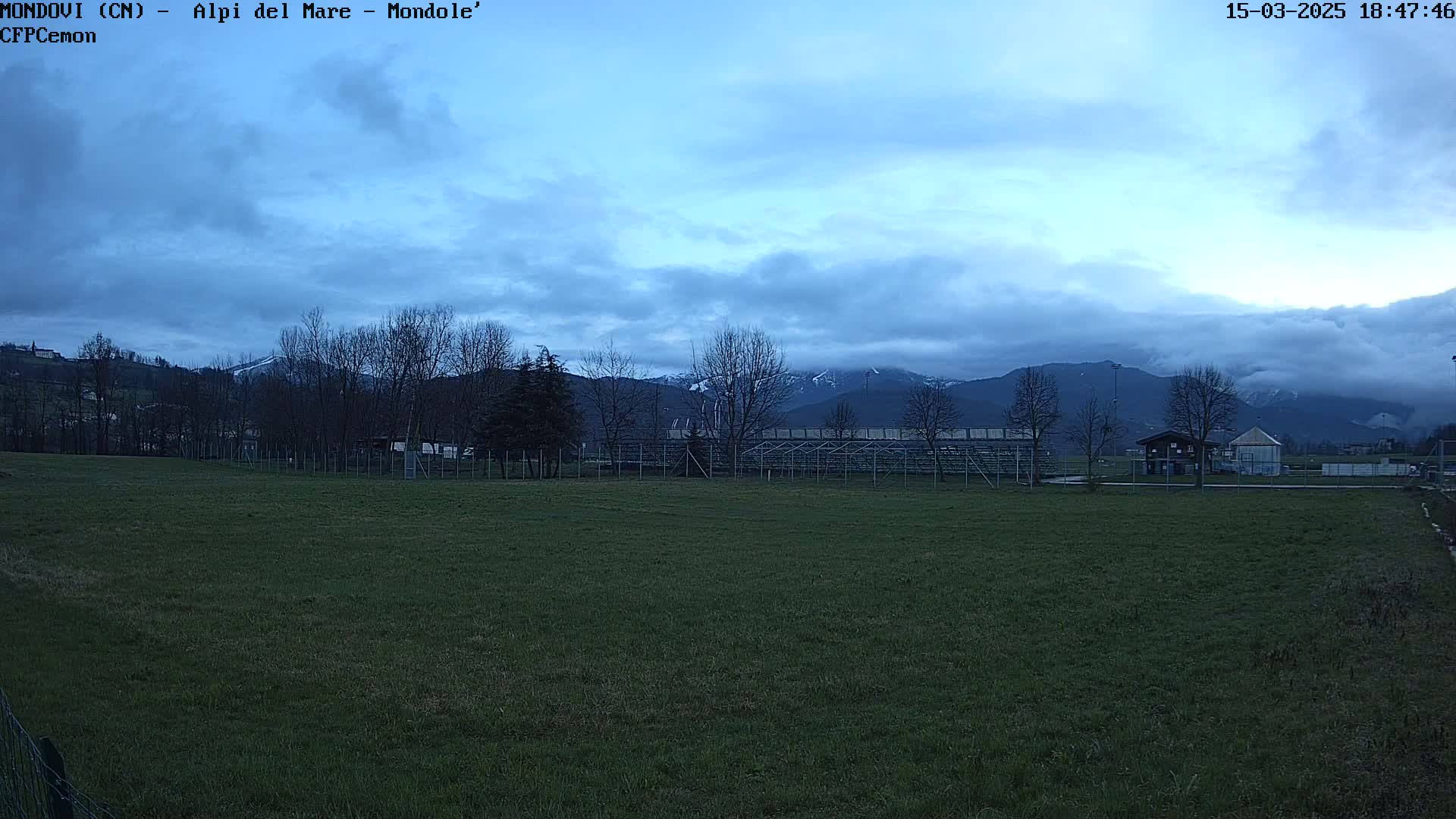 A grassy field with a bleacher seating area and bare trees sits in front of a mountain range under a mostly cloudy, dusky sky.