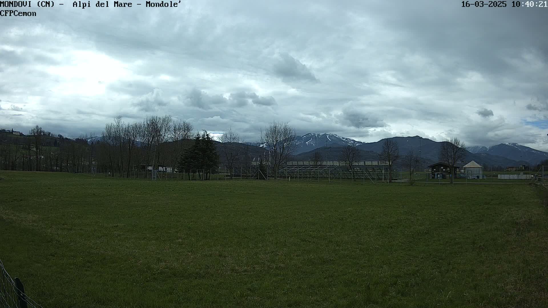 A grassy field with bleachers and a few buildings sits before a range of snow-capped mountains under a cloudy sky.