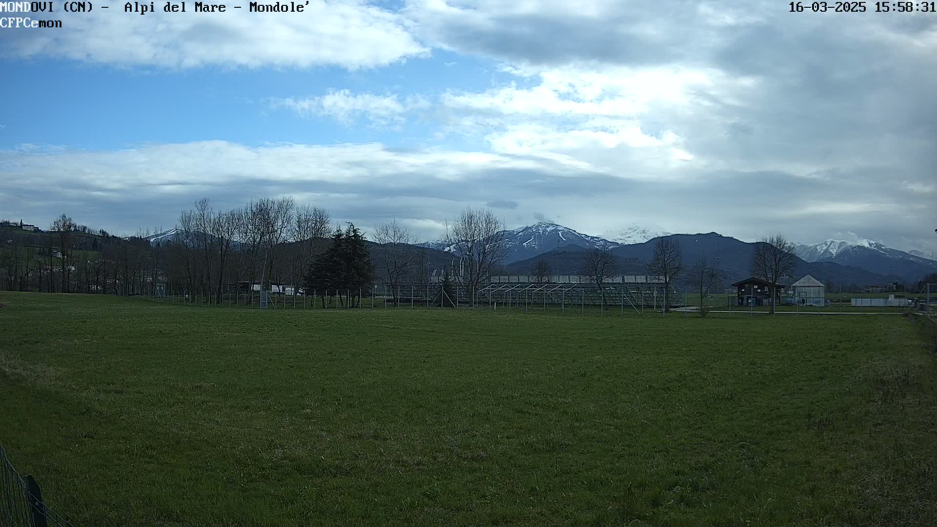 A grassy field with a few buildings and bleachers is situated in front of snow-capped mountains under a partly cloudy sky.