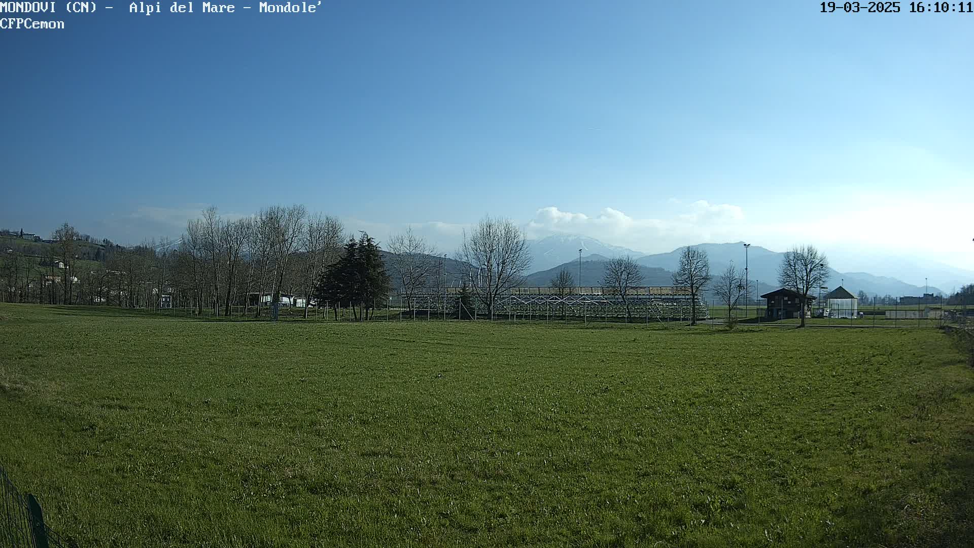 A grassy field stretches out before empty bleachers, with bare trees and distant mountains under a clear blue sky.