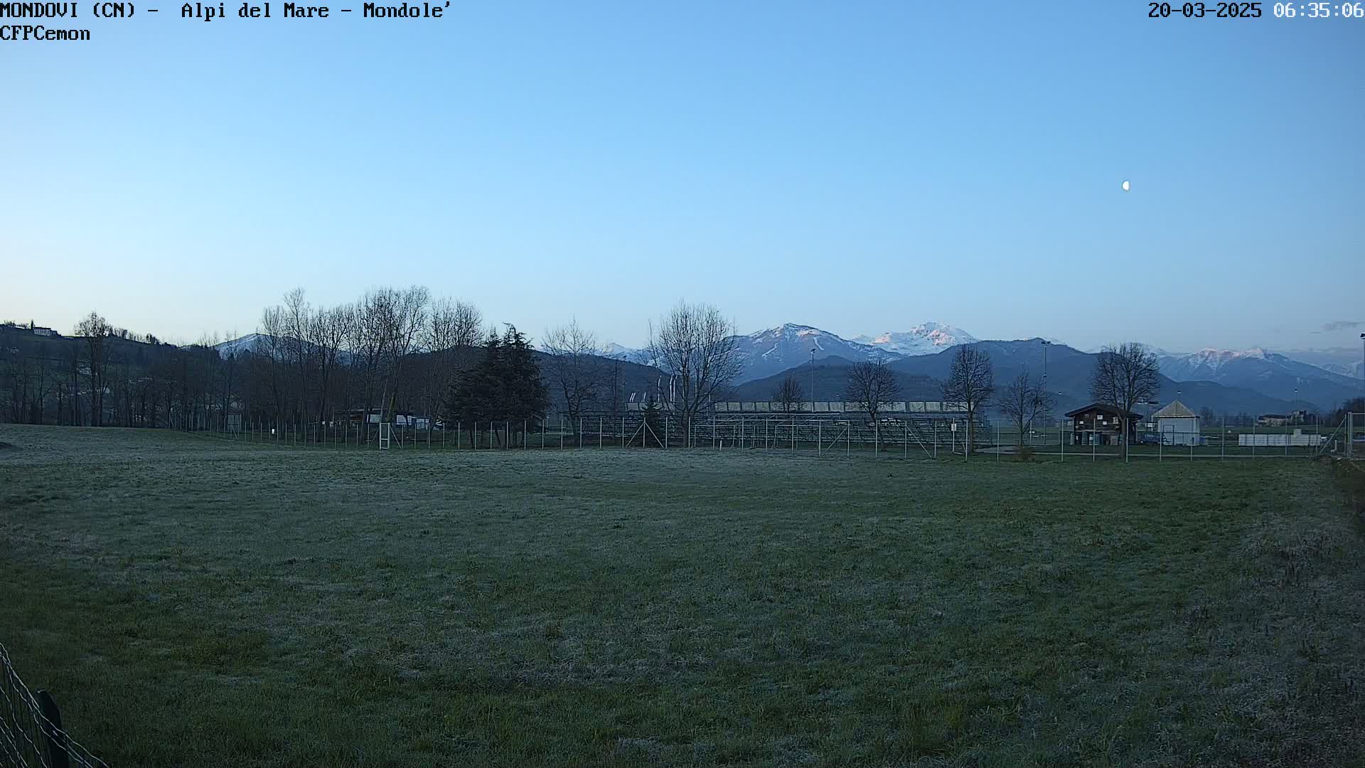 A grassy field with a bleacher structure and a small building sits before a snow-capped mountain range under a clear, twilight sky with a visible moon.