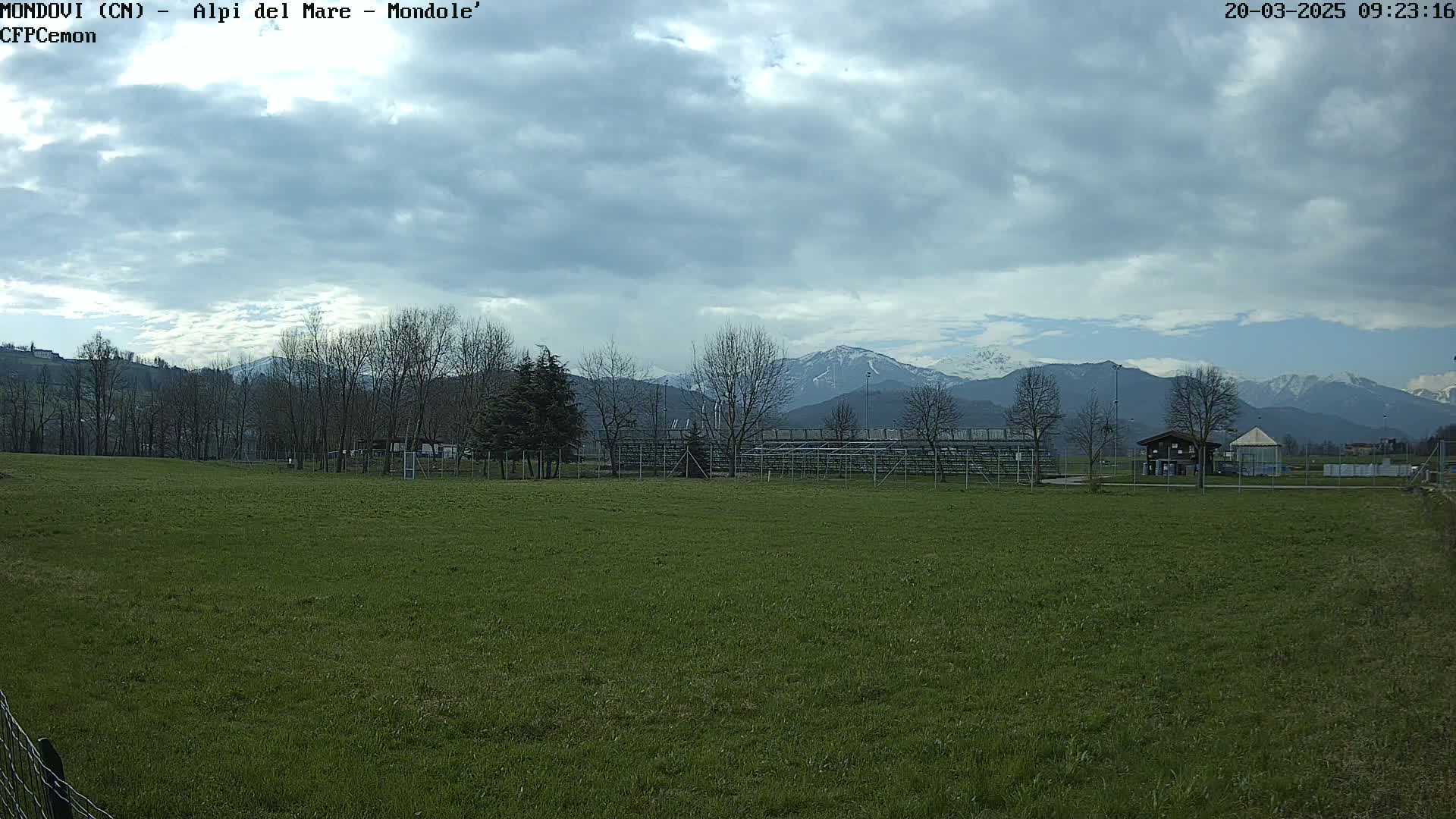 A grassy field with a few buildings and trees in the foreground, overlooks snow-capped mountains under a partly cloudy sky.