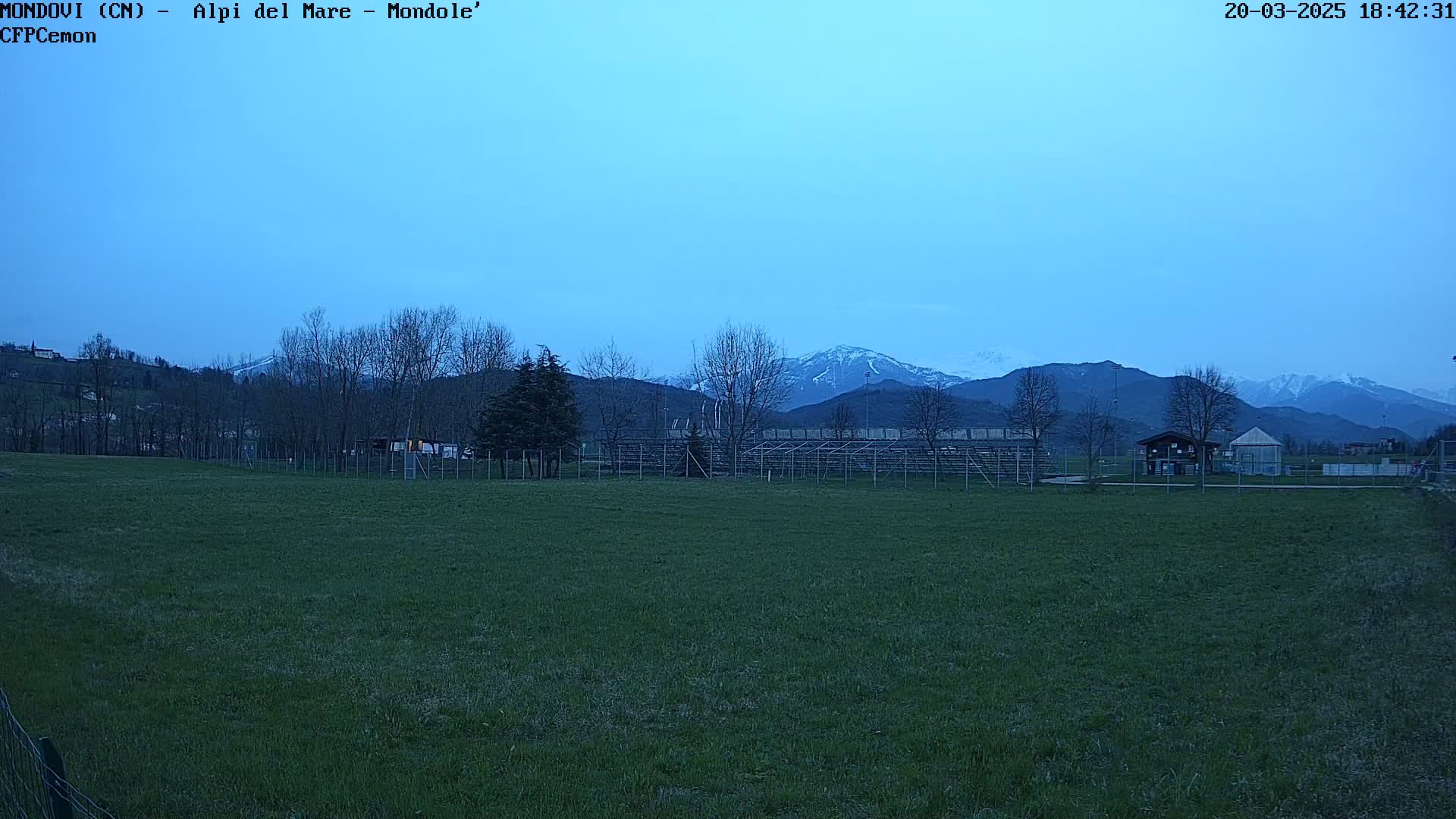A grassy field with bleachers and a small building sits in the foreground, under a twilight sky, with snow-capped mountains in the background.