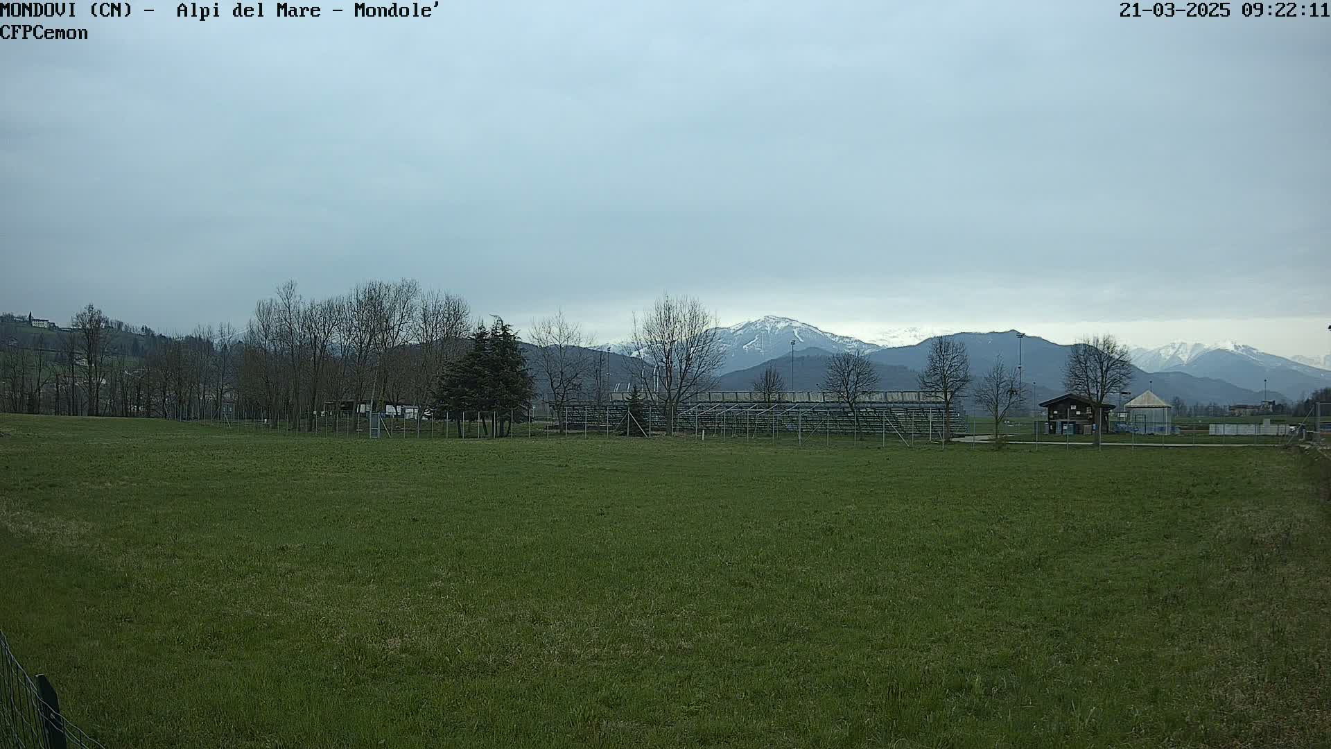 A grassy field with bleachers and a few small buildings stretches toward snow-capped mountains under a cloudy sky.