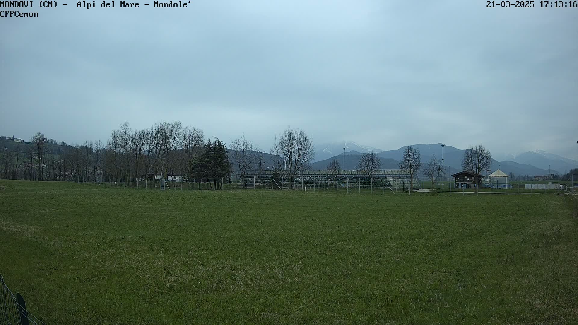 A grassy field with bleachers and a small building sits in front of bare trees and snow-capped mountains under a cloudy sky.