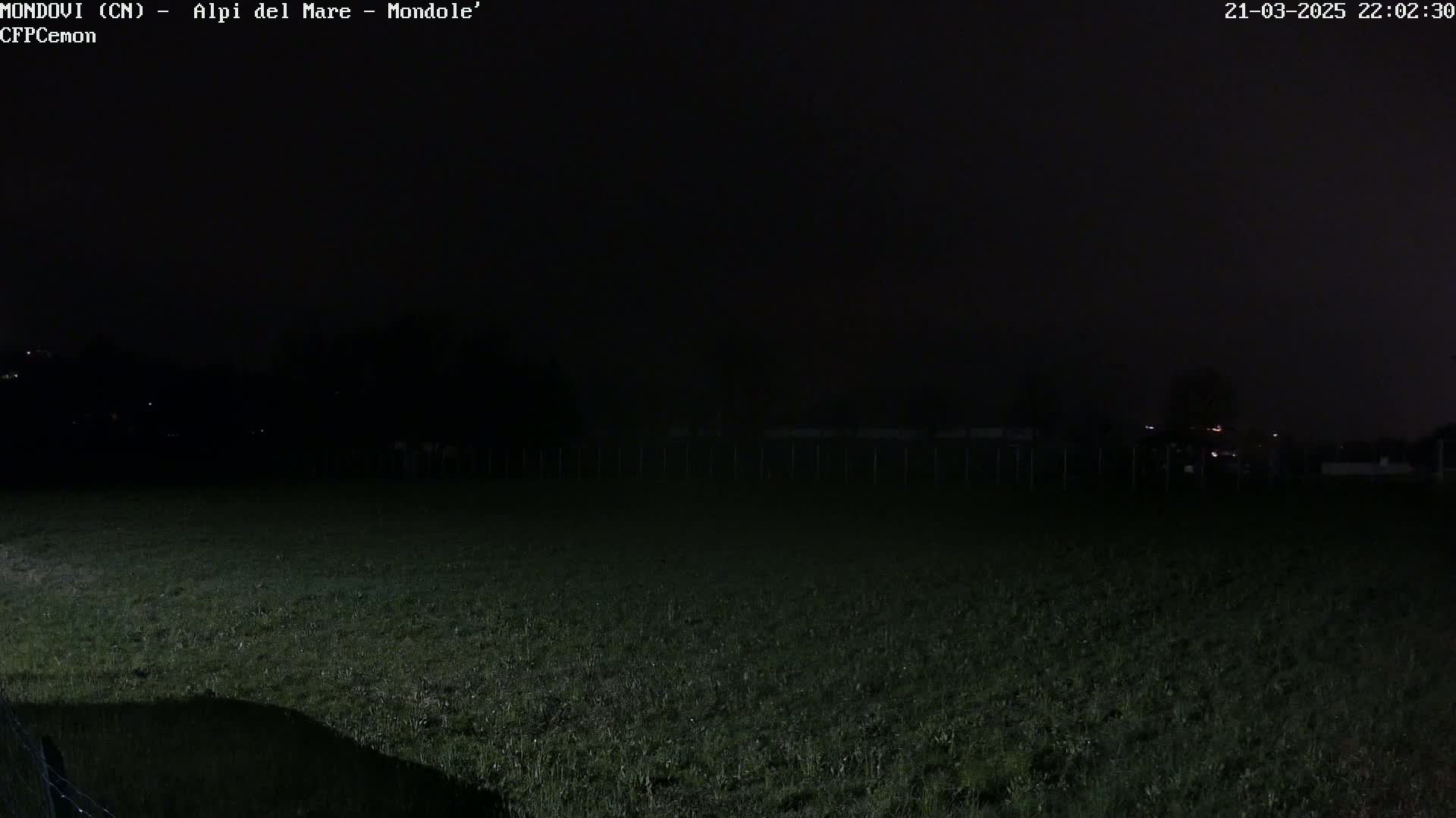 A dark, nighttime scene shows a grassy field with a fence and a small body of water, under a mostly cloudy sky.