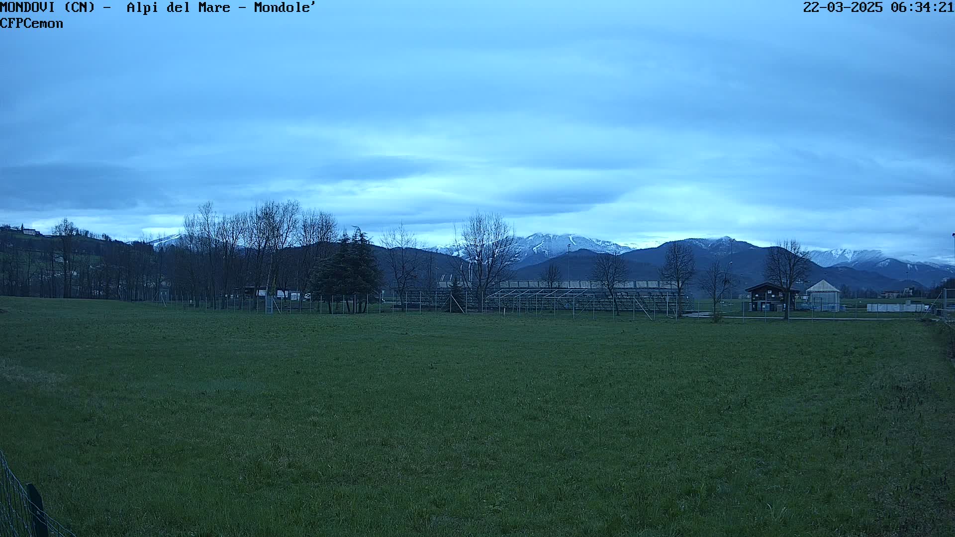 A grassy field with bleachers and a small building sits in the foreground, overlooking snow-capped mountains under a mostly cloudy, dusky blue sky.