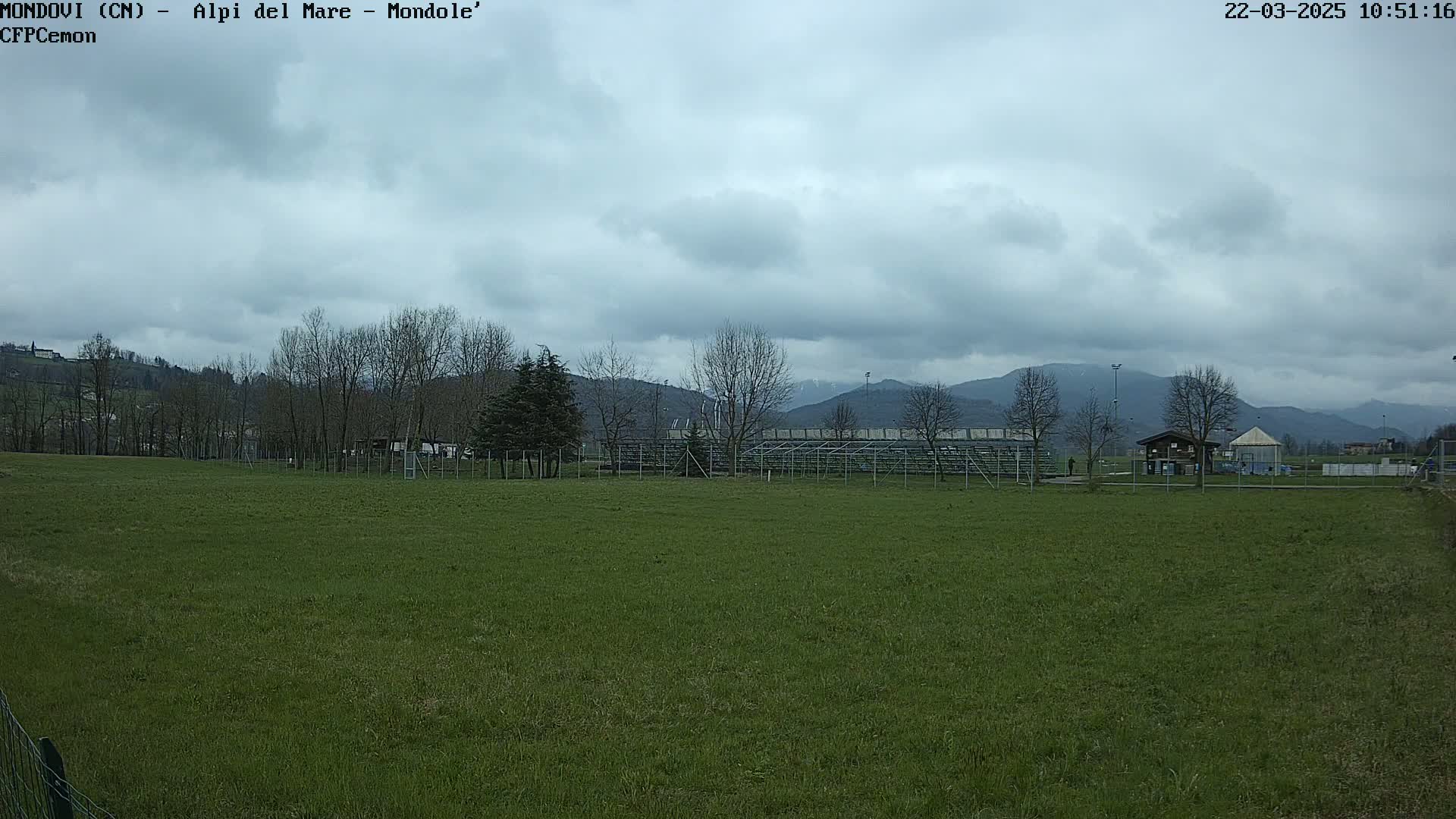 A grassy field with bleachers and a small building in the background under a cloudy sky.