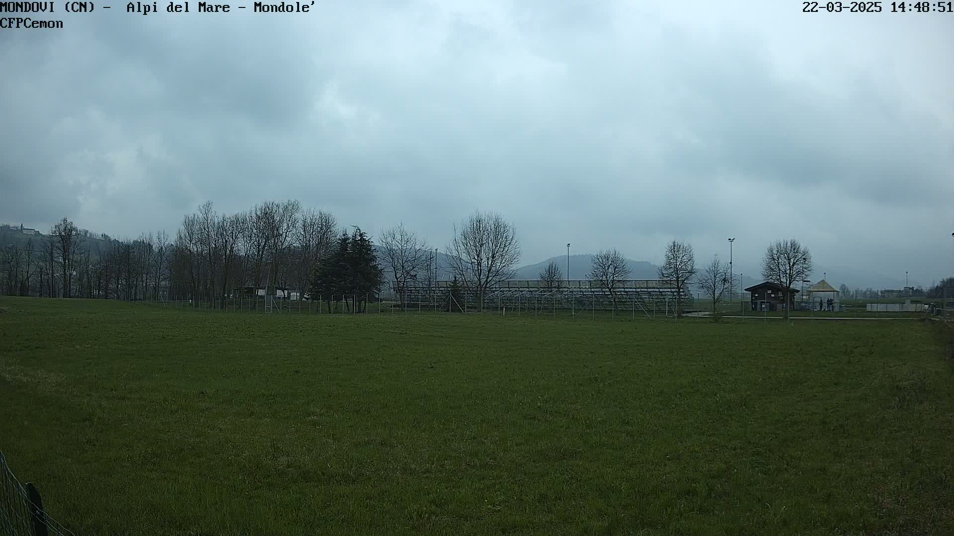 A grassy field with bleachers and a small building in the background under a cloudy sky.