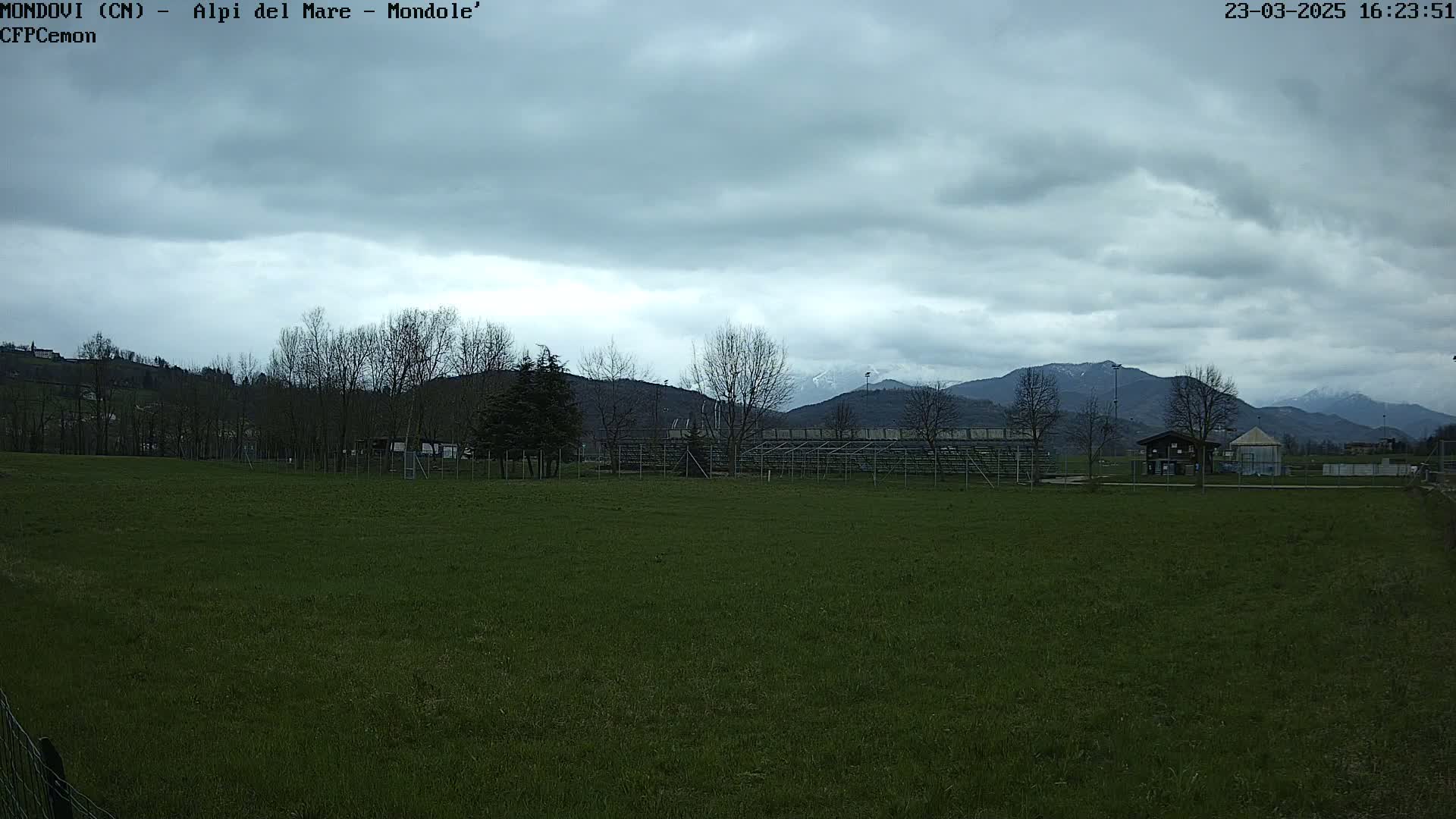 A grassy field sits before empty bleachers and a small building, with bare trees and snow-capped mountains in the background under an overcast sky.
