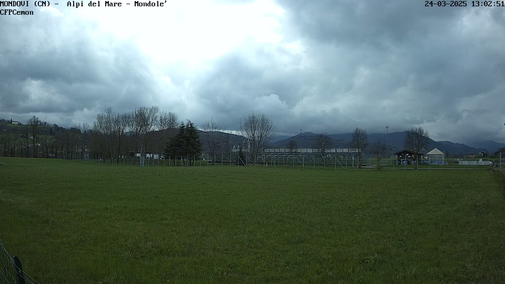 A grassy field stretches out before a metal bleacher structure, beyond which are bare trees and distant mountains under a cloudy sky.