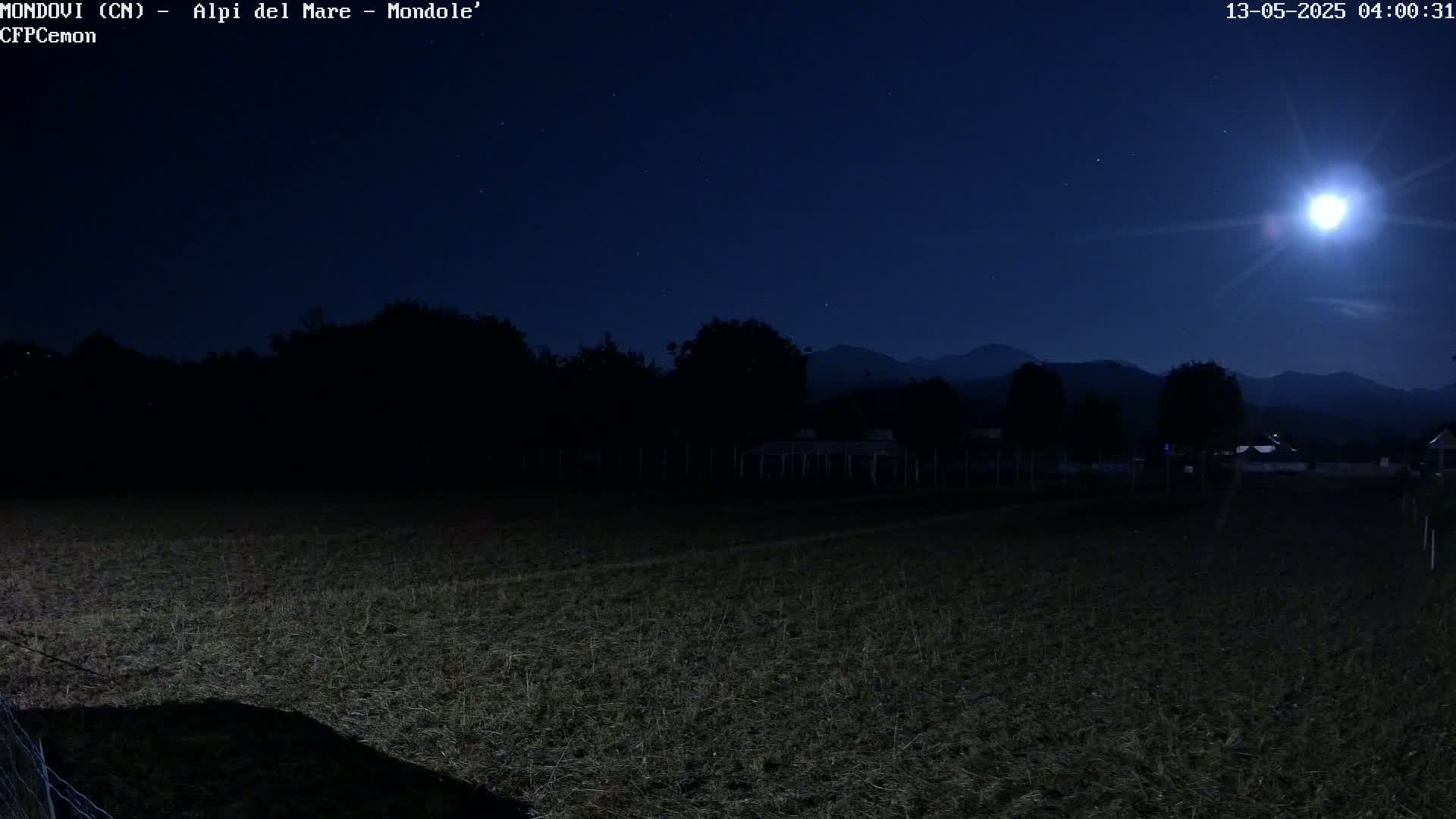 A bright moon illuminates a dark field and distant mountains under a mostly clear night sky.
