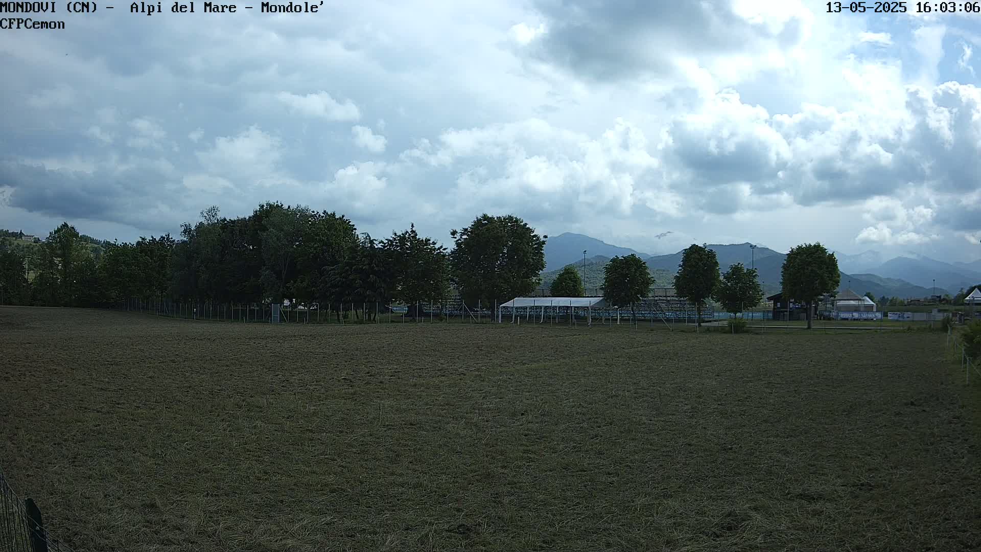 A mostly cloudy day reveals a recently mowed field, trees, a covered structure, and distant mountains.