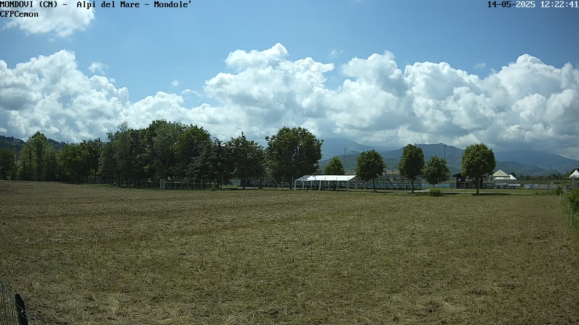 A mostly sunny day with puffy white clouds over a field of dry grass, beyond which are trees, a covered structure, and distant mountains.