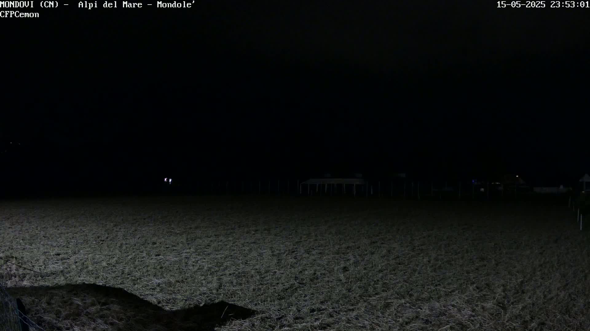 A dark, nighttime view of a field with some distant lights and a structure in the background.