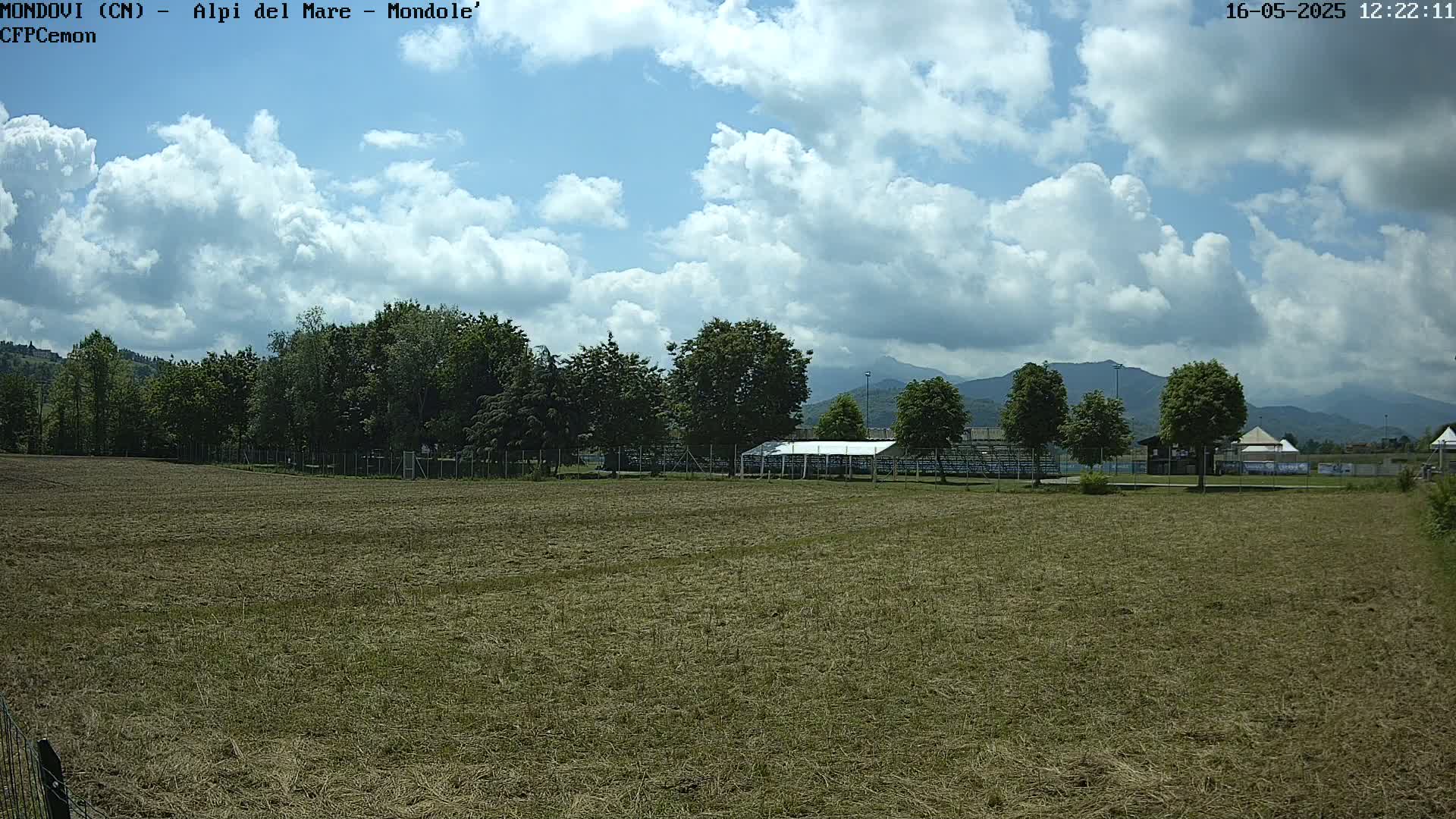 A mostly sunny day with puffy clouds over a mown field, behind which are trees, a covered structure, and distant mountains.
