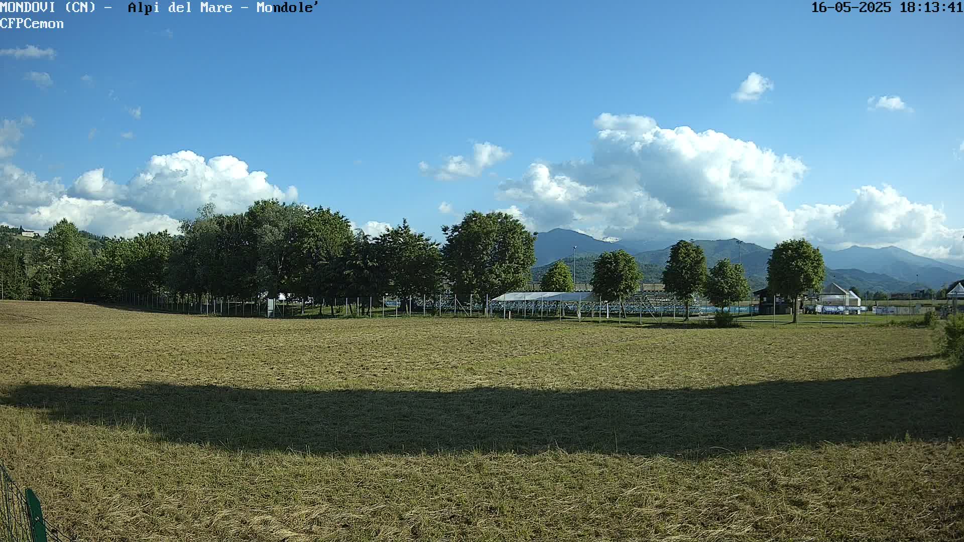 A mostly sunny day shows a grassy field, a line of trees, a covered structure, and mountains in the background.