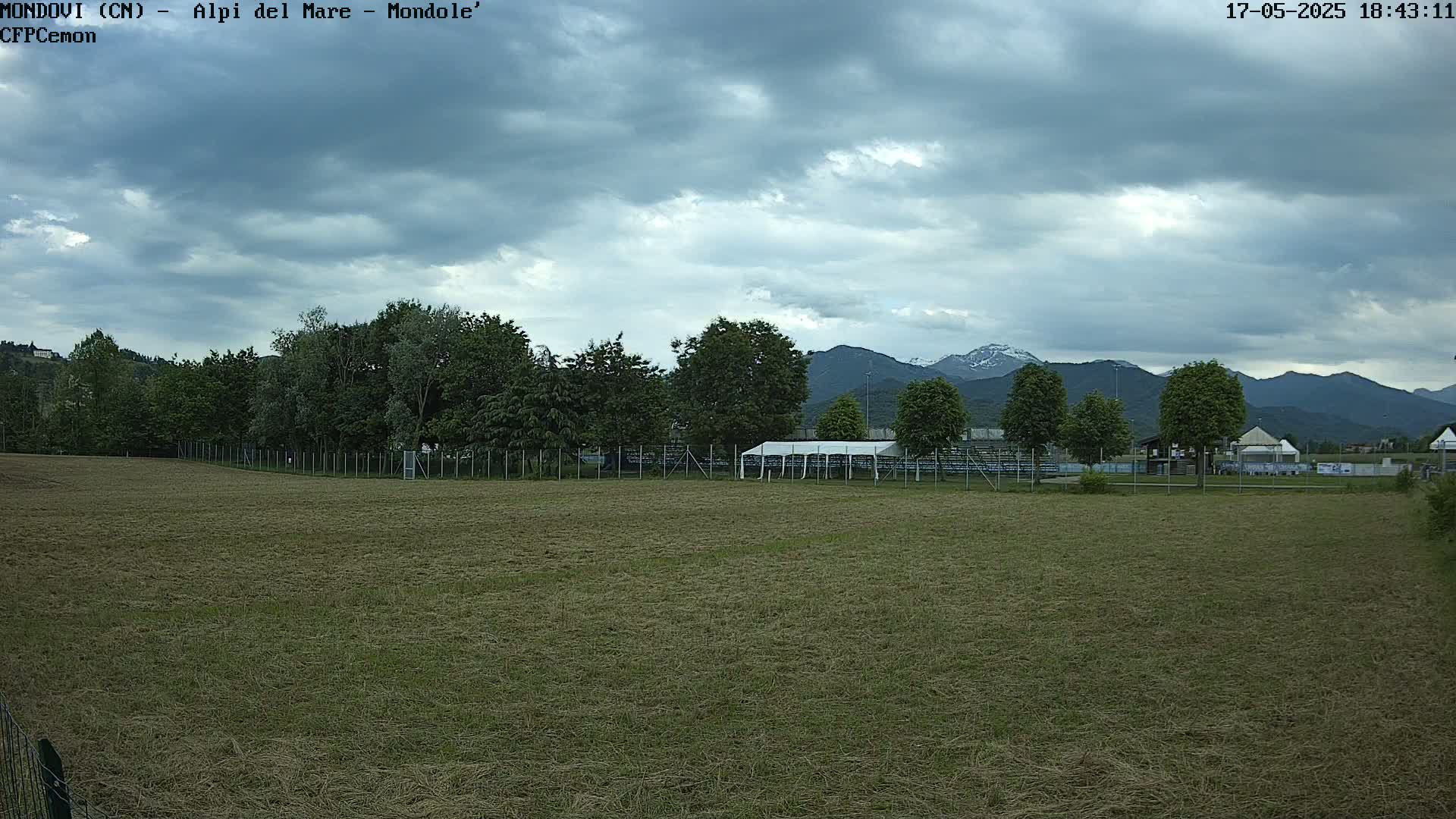 A grassy field, partially mowed, extends to a line of trees and a fenced-in area with a white tent, beyond which mountains are visible under a cloudy sky.