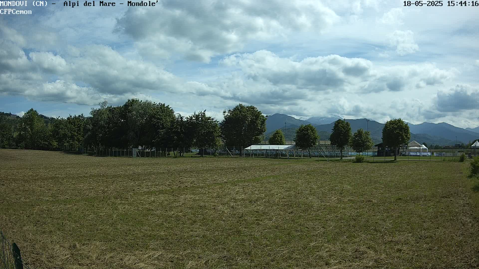 A mostly sunny day with partly cloudy skies shows a grassy field, trees, and bleachers in front of distant mountains.