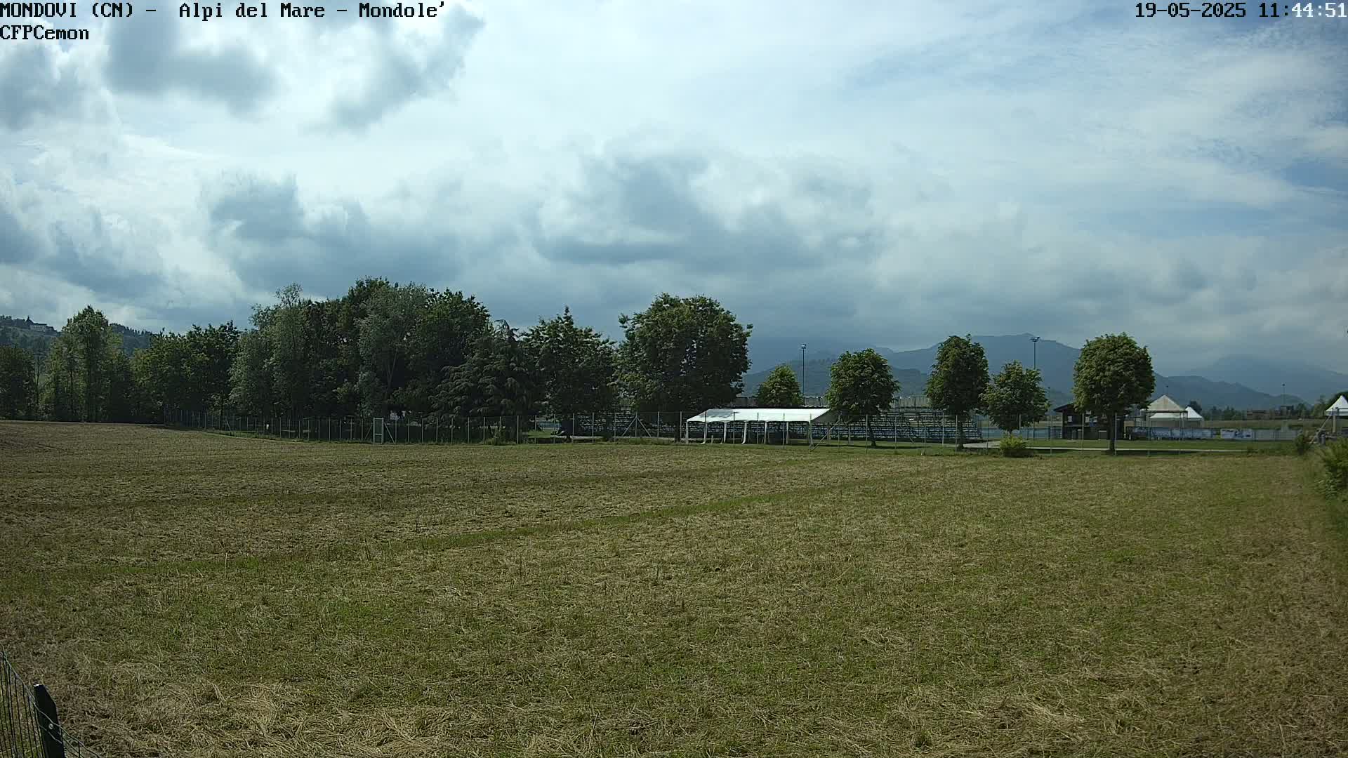 A mostly cloudy sky hangs over a mown field with trees and a white tent behind a fence, mountains visible in the distance.