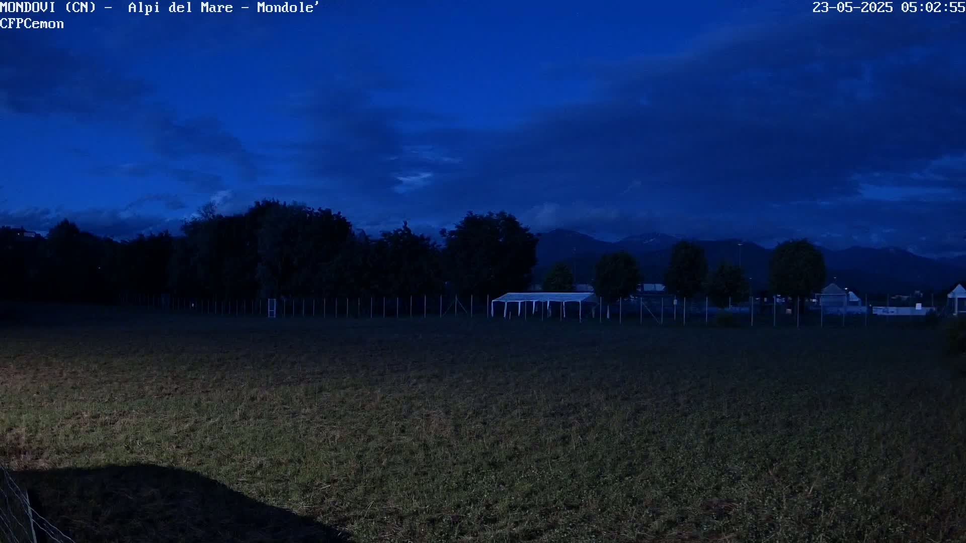 A dark, twilight scene shows a field with a white tent near a fence, trees, and mountains in the background under a mostly cloudy sky.