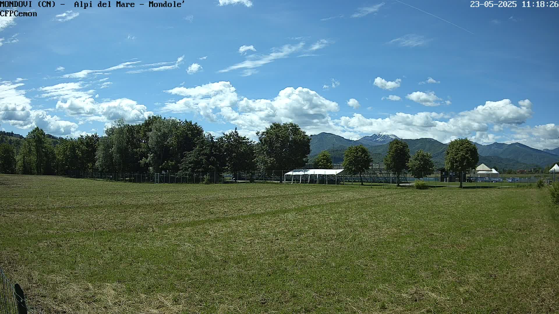 A grassy field with a white tent and trees in the background, under a partly cloudy sky and mountains in the distance.