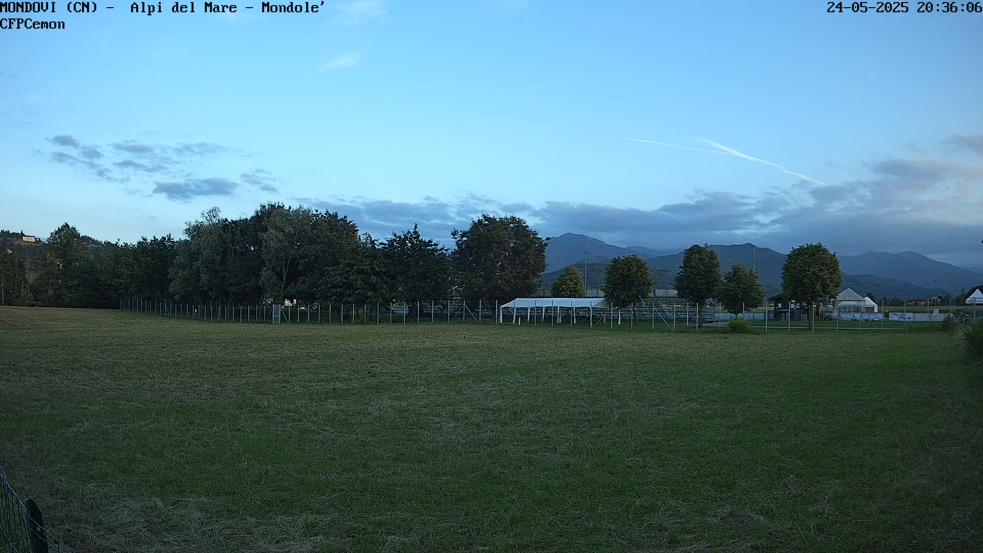 A grassy field with a white tent and bleachers behind a fence, under a mostly clear twilight sky with mountains in the background.