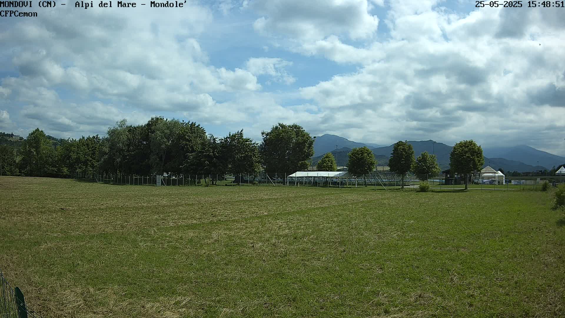 A partly cloudy sky overlooks a grassy field with trees, a covered structure, and distant mountains.