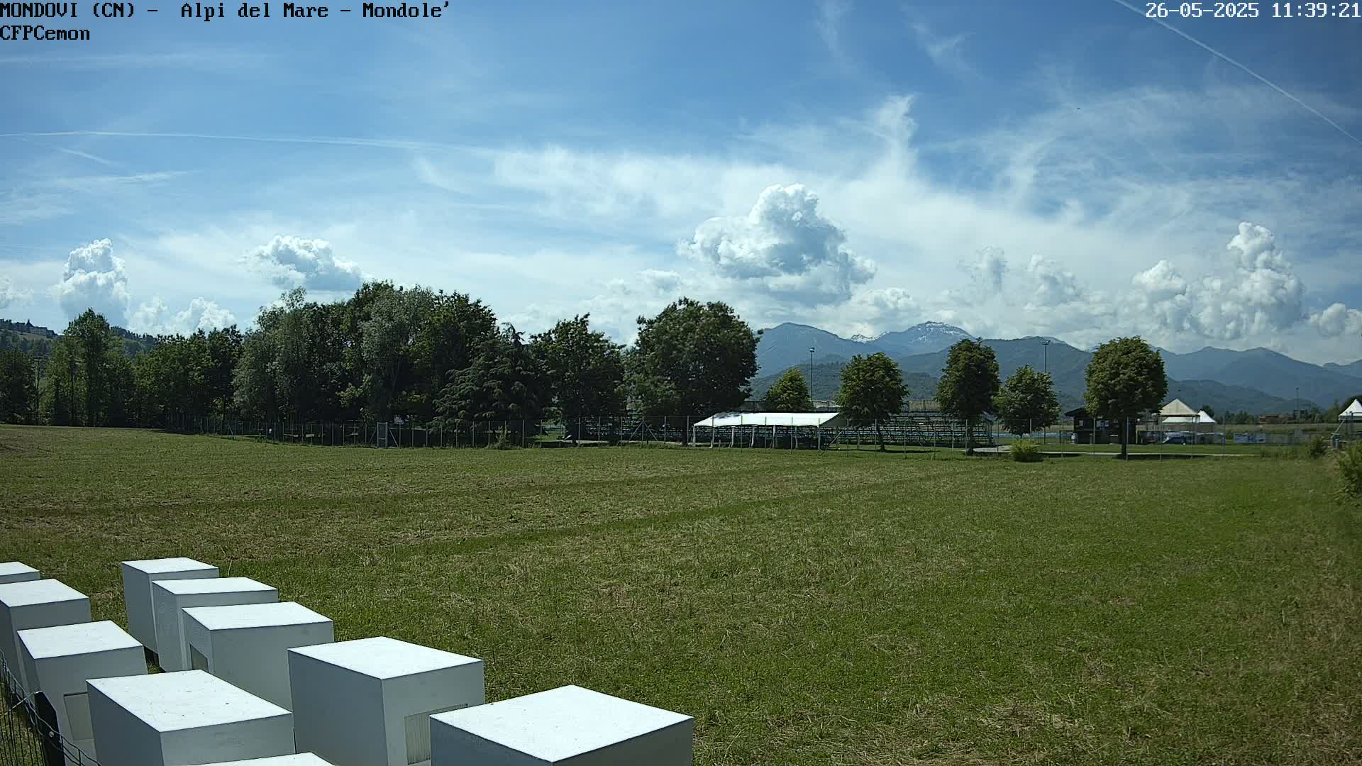 A grassy field with white cubic structures in the foreground, trees and a white tent in the middle ground, and mountains under a partly cloudy sky.