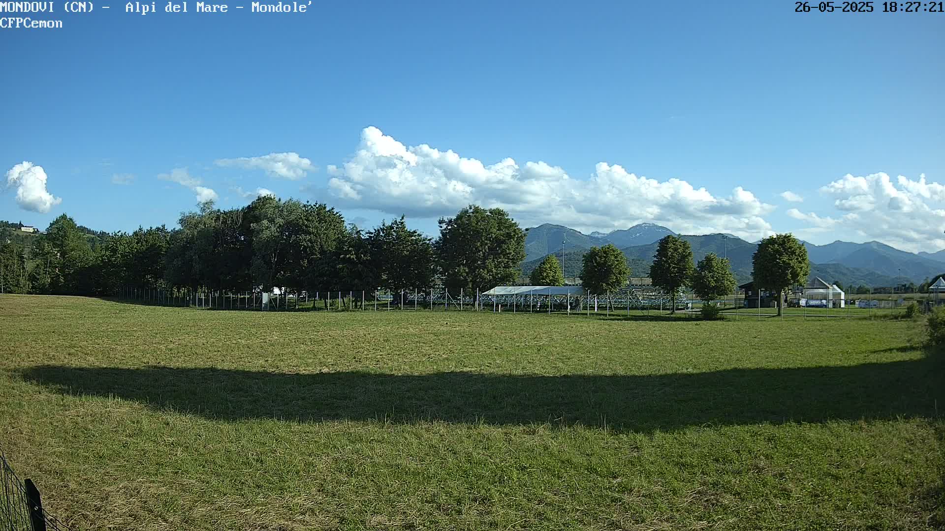 A grassy field stretches out before a line of trees, a covered structure, and distant mountains under a partly cloudy blue sky.