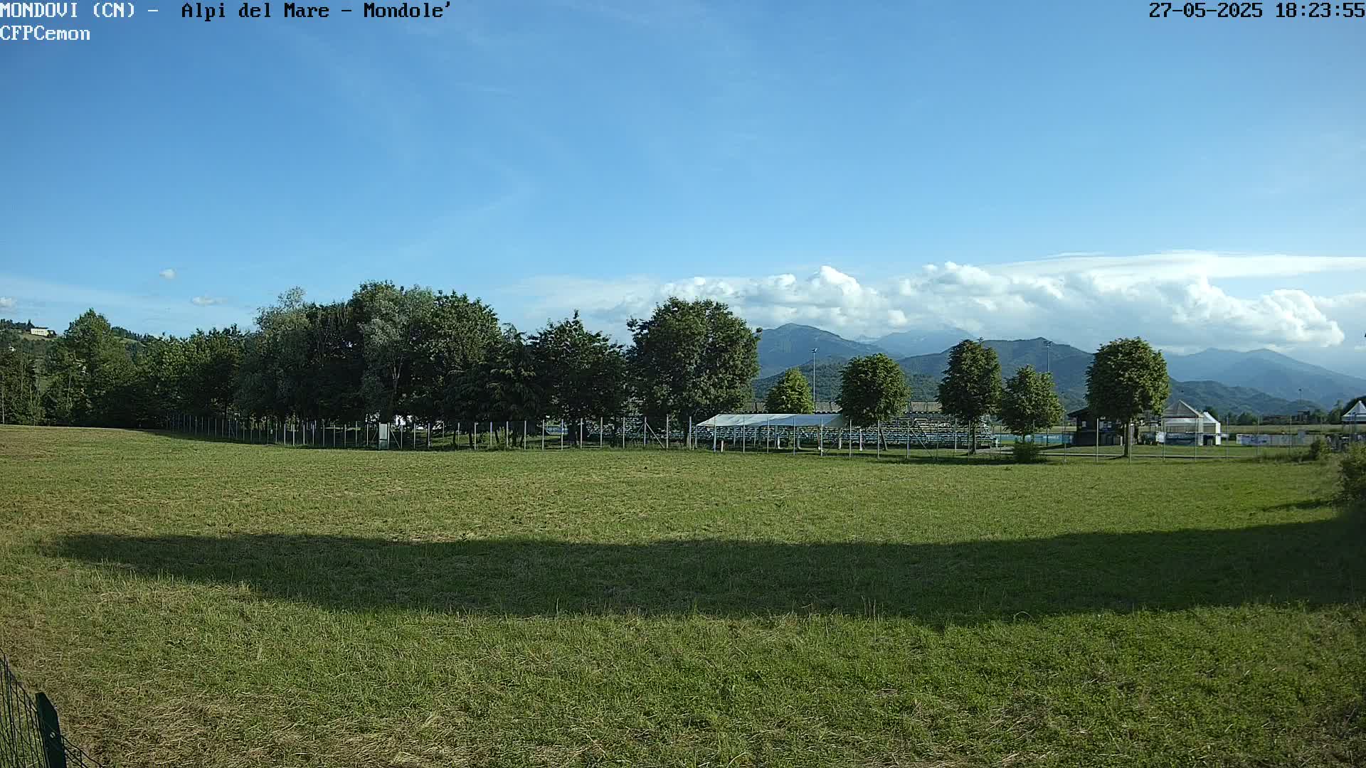 A grassy field with a line of trees and a covered structure behind it, mountains in the background under a mostly clear blue sky with some clouds.
