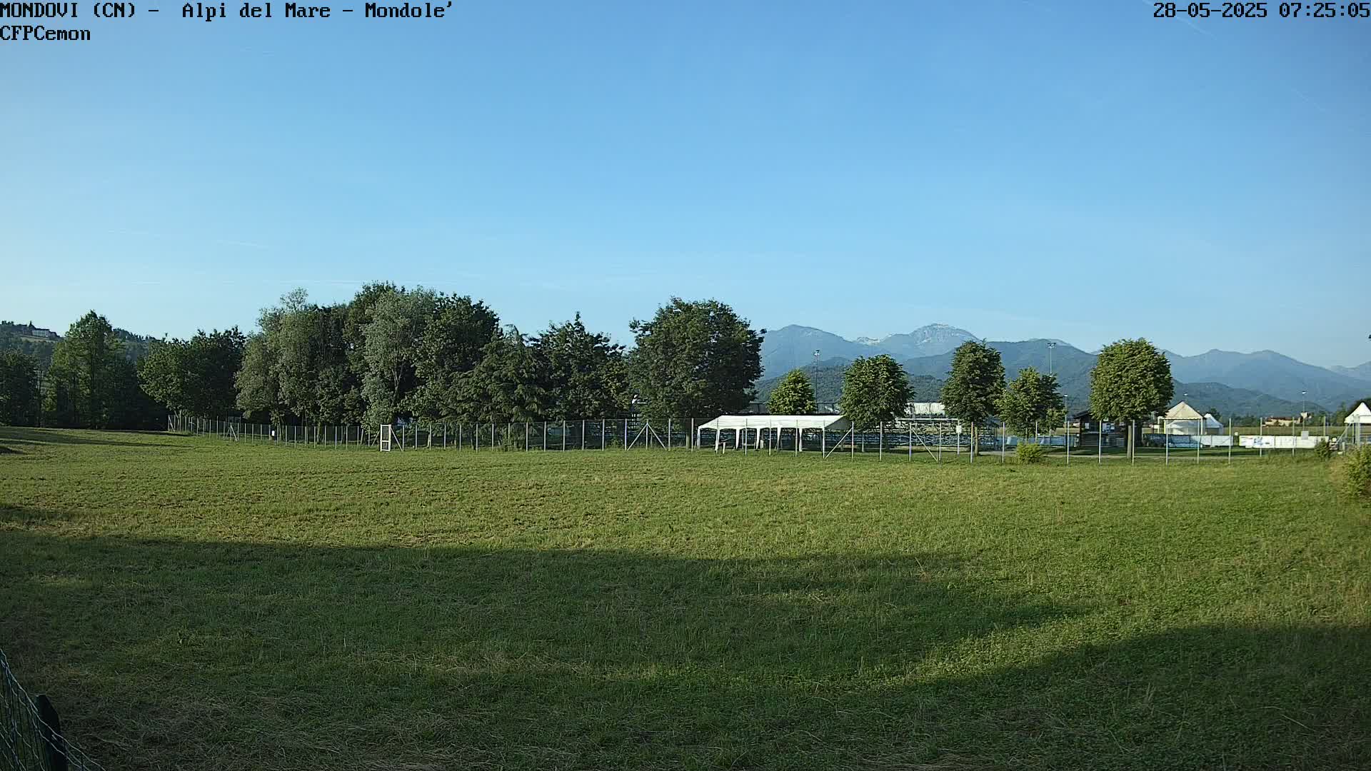 A grassy field with a line of trees and a white tent behind a fence, mountains in the background under a clear blue sky.