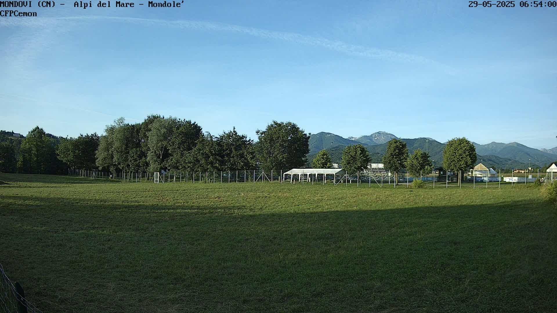 A grassy field enclosed by a fence, with trees and a white pavilion in the background, under a clear blue sky and mountains in the distance.