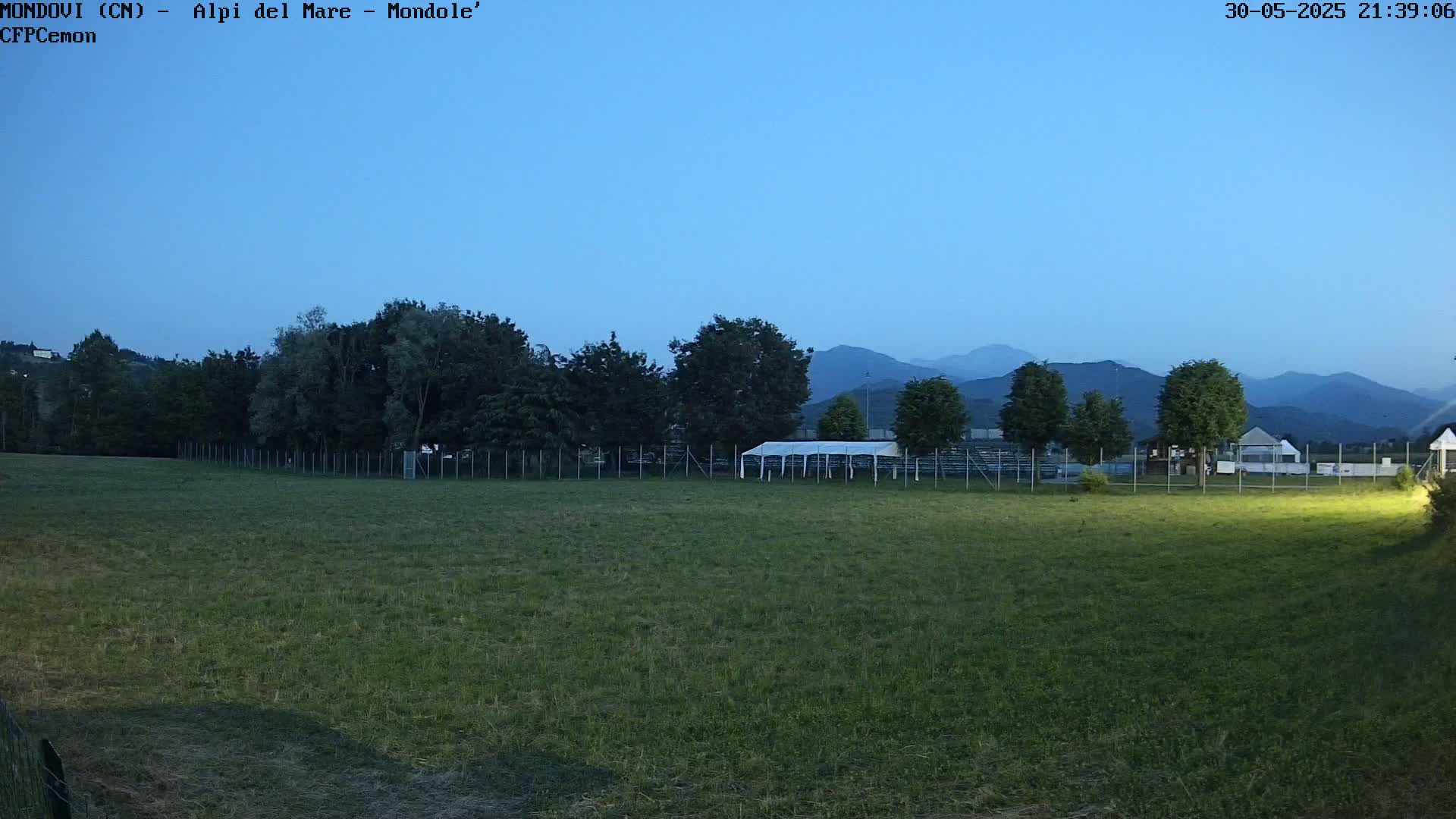 A large grassy field at dusk is enclosed by a fence, behind which are some trees and a white tent, with mountains visible in the background under a clear twilight sky.
