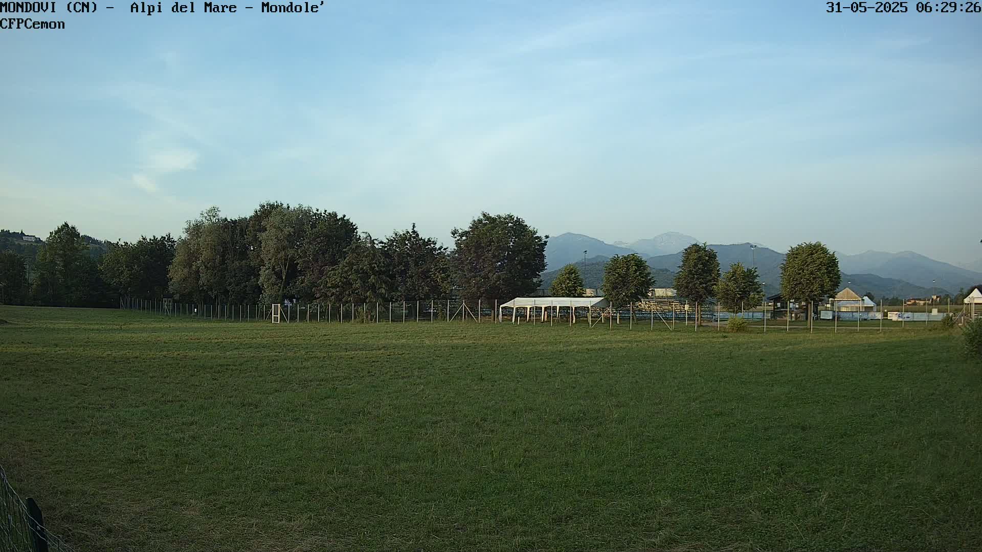 A grassy field with a fence, trees, and a white tent in the foreground, mountains in the background under a mostly clear sky.