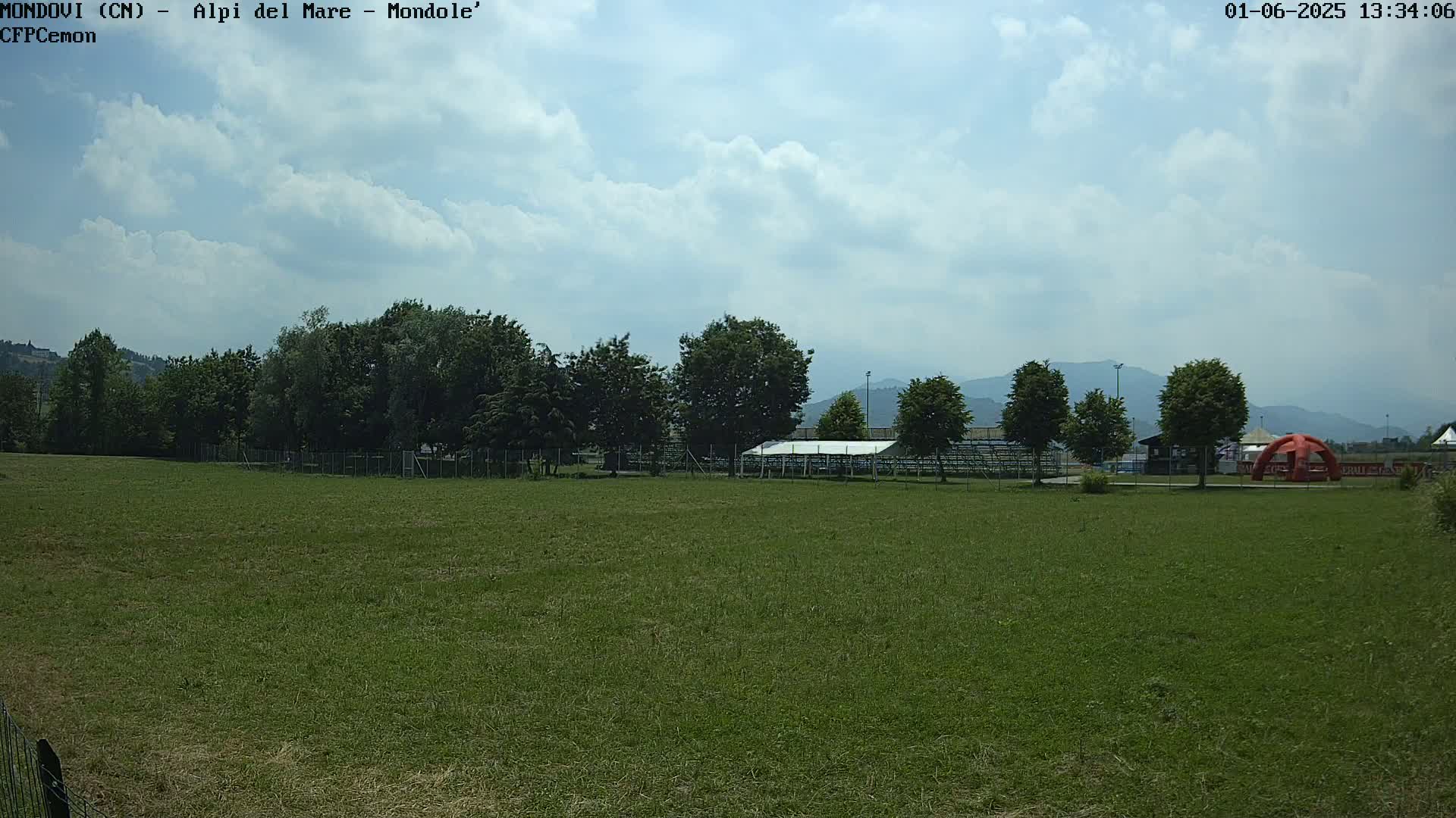 A large grassy field stretches towards a line of trees and a partially visible structure under a partly cloudy sky.