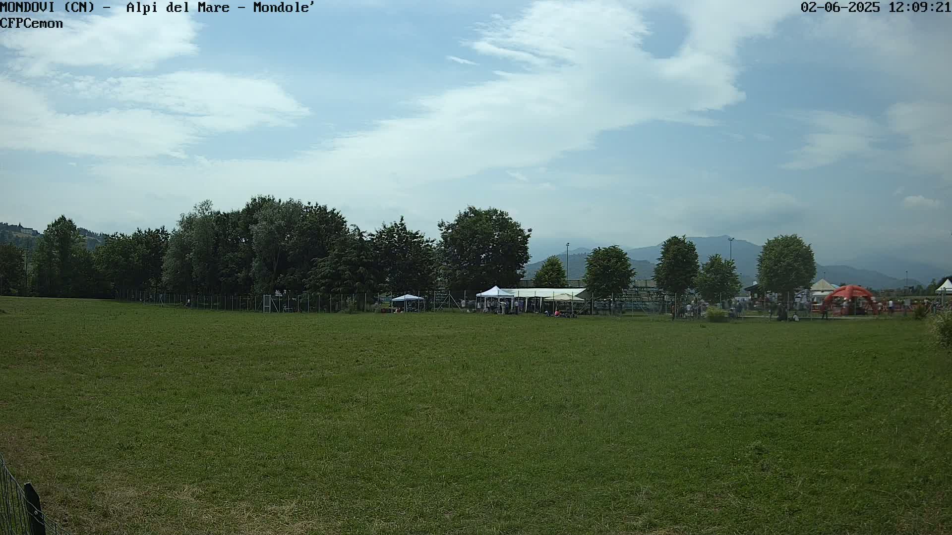 A grassy field with a few people and tents in the background under a partly cloudy sky, with mountains visible in the distance.