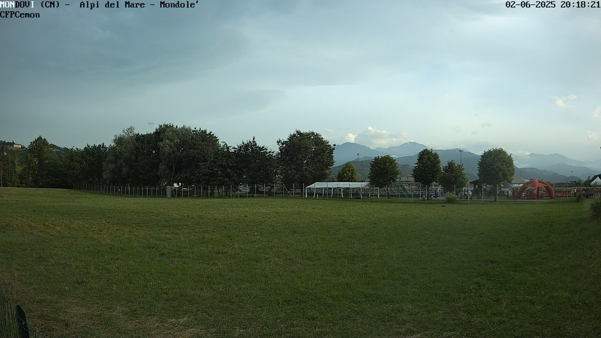 A grassy field stretches out before a line of trees and bleachers, with hazy mountains in the background under a partly cloudy sky.