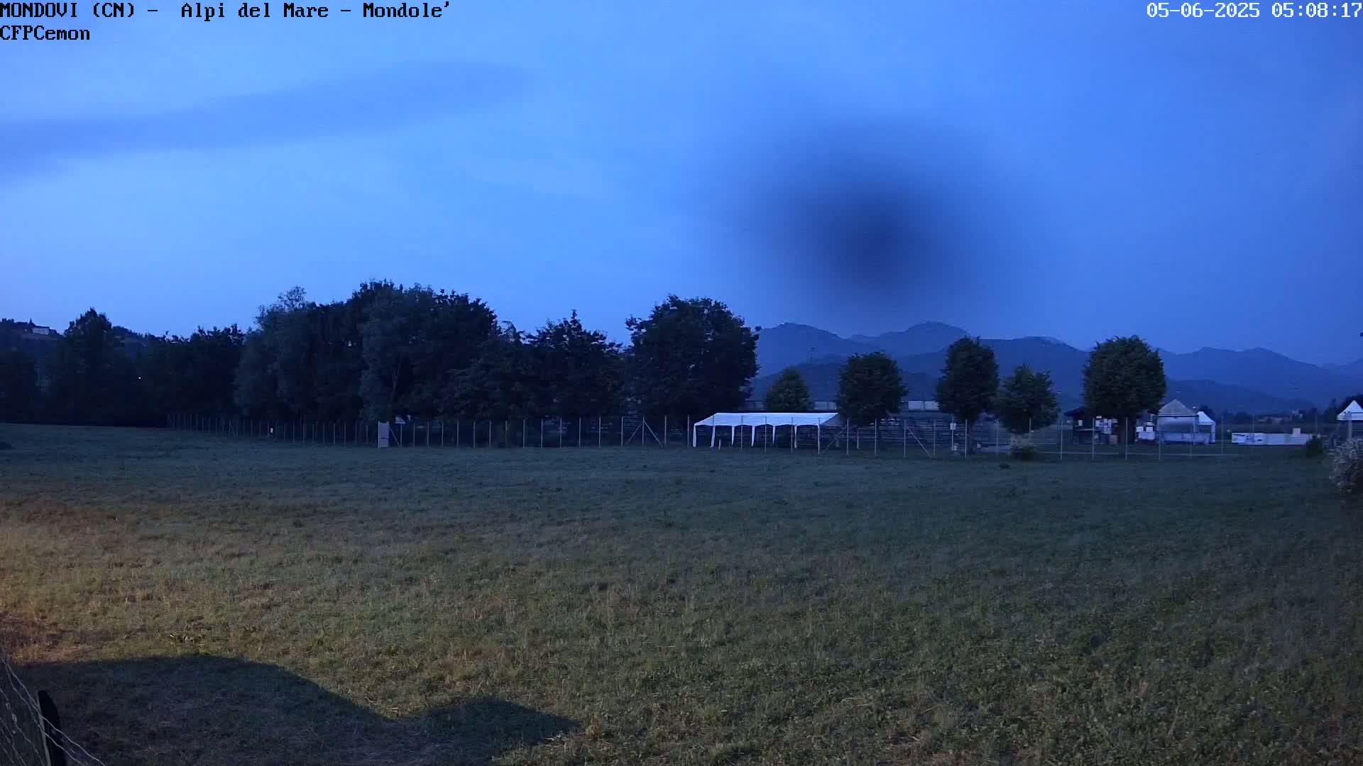 A twilight view of a grassy field enclosed by a fence, with trees, a large white tent, and distant mountains under a clear, dark-blue sky.