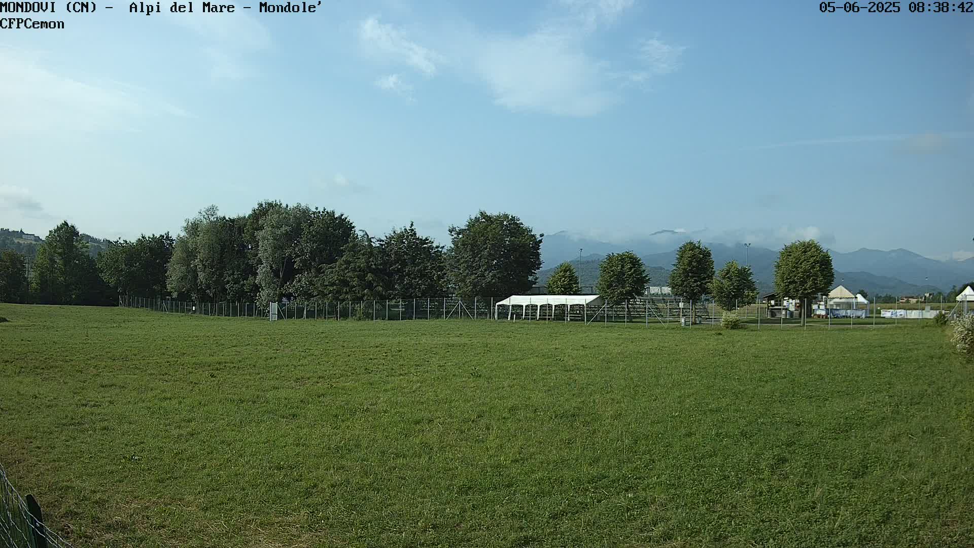 A large grassy field stretches out before a line of trees, beyond which are mountains partially obscured by haze under a mostly clear sky.