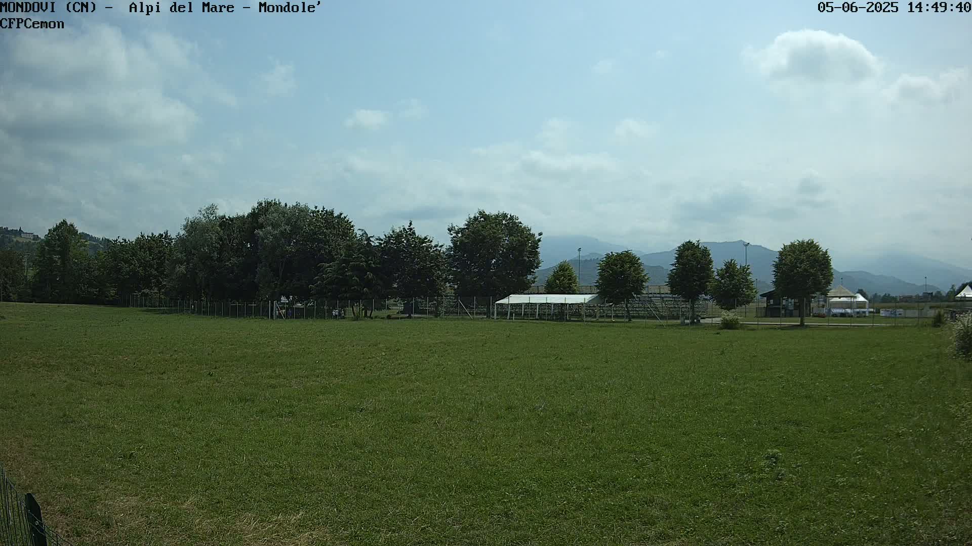 A mostly sunny day reveals a grassy field with trees, a small structure, and bleachers in the background, mountains visible in the distance.
