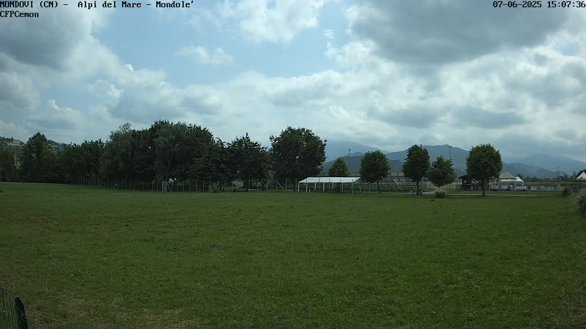 A large grassy field stretches out under a partly cloudy sky, with trees and a white canopy visible in the midground, and mountains in the far distance.