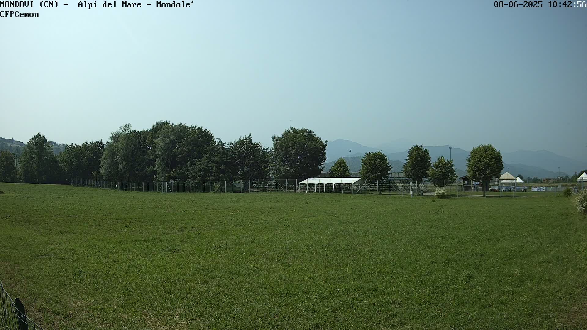 A large grassy field stretches out before a line of trees and a white tent structure, all set against a backdrop of distant mountains under a clear, sunny sky.